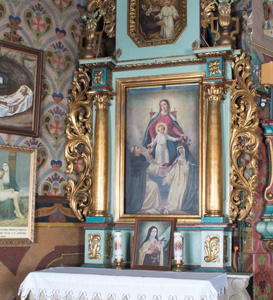 Zdjęcie nr 2: Obraz w formie stojącego prostokąta z przedstawieniem tronującej Matki Boskiej z Dzieciątkiem i klęczącymi przed nią świętymi dominikańskimi. Maria siedzi na tronie unoszącym się wśród obłoków, zwrócona jest frontalnie, z głową skierowaną w prawo. Prawą ręką trzyma za ramię stojącego przed nią Jezusa, w lewej trzyma wieniec z róż nad głową św. Katarzyny. Twarz ma owalną, o łagodnych rysach, oczach skierowanych w dół, prostym nosie i małych ustach, okoloną brązowymi włosami, schowanymi pod welonem. Ubrana jest w ciemnoróżową suknię, niebieski płaszcz ze złoconą lamówką, zapięty pod szyją, zarzucony na ramiona i opadający na kolana, na głowie ma biały welon. Dzieciątko w pozycji stojącej, w kontrapoście, zwrócone jest frontalnie, z głową skierowaną w prawo. Lewą rękę trzyma nad głową św. Katarzyny, prawą podaje różaniec św. Dominikowi. Twarz okrągła, o pełnych policzkach, dużych oczach, małym nosie i pełnych ustach, okolona jasnymi, krótkimi, kręconymi włosami. Jezus ubrany jest w białą tunikę przepasaną w talii. Wokół głów Marii i Dzieciątka nimby promieniste. Po prawej stronie Marii klęczy na jedno kolano św. Dominik. Ukazany jest z profilu, z uniesioną głową i rozłożonymi na boki rękoma, w których podtrzymuje różaniec. Twarz ma szczupłą, z oczami skierowanymi ku górze i z tonsurą na głowie. Ubrany jest w biały habit i czarny płaszcz z kapturem. Po lewej stronie Marii klęczy św. Katarzyna Sieneńska zwrócona w trzech czwartych w prawo, z pochyloną głową. W lewej, uniesionej ręce trzyma różaniec, prawą wyciąga w bok. Twarz ma o łagodnych rysach, zwróconych w dół oczach, prostym nosie i pełnych ustach. Ubrana jest w biały habit ze szkaplerzem oraz czarny płaszcz i welon. Na głowie ma koronę cierniową. Pomiędzy świętymi leży otwarta księga i gałązka lilii. 