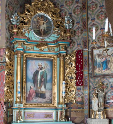 Zdjęcie nr 1: Nastawa ołtarzowa architektoniczna, jednoosiowa, jednokondygnacyjna ze zwieńczeniem ustawiona na cokole z wyodrębnionymi po bokach prostopadłościanami. Ołtarz kamienny prostopadłościenny, ujęty po bokach esownicami, z przodu flankowany kanelowanymi lizenami, w centrum krzyż. Nastawa flankowana parą kolumn o kapitelach kompozytowych, dźwigających pełne belkowanie wyłamane na ich osi, w partii fryzu dekorowane rozetami, wicią roślinną i muszlą. Całe pole środkowe wypełnia nisza w formie stojącego prostokąta z obrazem św. Wojciecha, ujęta złoconą ramą zdobioną dekoracją roślinną. Na belkowaniu ustawione dwa wazony z bukietami kwiatów. Kondygnacja flankowana uszami utworzonymi z ażurowego akantu. Zwieńczenie w formie owalnego pola z obrazem Męczeństwa św. Sebastiana ujętego wieńcem laurowym i ażurowymi liśćmi akantu, u dołu dwa rogi obfitości. Całość zakończona medalionem z okiem Opatrzności w glorii promienistej. Struktura polichromowana w kolorze jasnoniebieskim, z niebieskimi i czerwonymi detalami, kolumny, profile i ornamentyka złocone, elementy dekoracji srebrzone. 