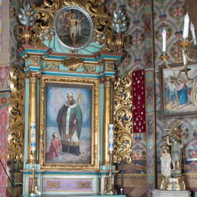 Zdjęcie nr 1: Nastawa ołtarzowa architektoniczna, jednoosiowa, jednokondygnacyjna ze zwieńczeniem ustawiona na cokole z wyodrębnionymi po bokach prostopadłościanami. Ołtarz kamienny prostopadłościenny, ujęty po bokach esownicami, z przodu flankowany kanelowanymi lizenami, w centrum krzyż. Nastawa flankowana parą kolumn o kapitelach kompozytowych, dźwigających pełne belkowanie wyłamane na ich osi, w partii fryzu dekorowane rozetami, wicią roślinną i muszlą. Całe pole środkowe wypełnia nisza w formie stojącego prostokąta z obrazem św. Wojciecha, ujęta złoconą ramą zdobioną dekoracją roślinną. Na belkowaniu ustawione dwa wazony z bukietami kwiatów. Kondygnacja flankowana uszami utworzonymi z ażurowego akantu. Zwieńczenie w formie owalnego pola z obrazem Męczeństwa św. Sebastiana ujętego wieńcem laurowym i ażurowymi liśćmi akantu, u dołu dwa rogi obfitości. Całość zakończona medalionem z okiem Opatrzności w glorii promienistej. Struktura polichromowana w kolorze jasnoniebieskim, z niebieskimi i czerwonymi detalami, kolumny, profile i ornamentyka złocone, elementy dekoracji srebrzone. 