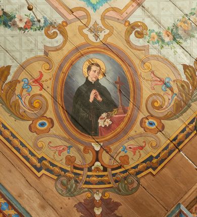 Zdjęcie nr 9: W prezbiterium w dolnej połowie ścian polichromia w formie namalowanej jasnoczerwonej, wielkowzorzytej tkaniny o motywach geometrycznych i roślinnych z niebieskim monogramem Marii i żółtymi koronami; jej górny brzeg odwiedziony jest pasem z brązowo-czerwoną dekoracją roślinną. Ściana przedzielona w połowie wysokości pasem z arkadkami wypełnionymi żółtymi wazonami oraz dekoracją roślinną na zielonym tle. W ich przyłuczach czerwone pola wypełnione malowaną roślinnością. W górnej połowie ścian malowane płyciny o jasnobrązowych ramach, których pola wypełnione są wzorem o motywie żółtych sześcioboków z fioletowymi kwiatami. Na południowej ścianie prezbiterium okna obwiedzione malowanymi, mazerowanymi ramami. W górnej części ściany, pod belkowaniem fryz utworzony z uskrzydlonych główek anielskich ukazanych na niebieskim tle, pomiędzy którymi znajdują się czerwone kampanule, pod nim podwieszone kwiatowe girlandy. Ściany zakończone profilowanym gzymsem, pomalowanym w dwa pasy – czerwony i niebieski, dekorowane motywem liści. Ściany podzielone lizenami w kolorze fioletowym, zdobionymi motywami geometrycznymi, różnymi w dolnej i górnej połowie. Strop prezbiterium z namalowaną płyciną w formie stojącego prostokąta, zamkniętego u dołu trójbocznie, o wklęsłych sześciu narożach, w których namalowana jest dekoracja roślinna. Płycina ujęta brązowo-żółtą ramą zdobioną motywem roślinnym oraz niebieskim pasem, z abstrakcyjnym motywem na wklęśnięciach. Różowe pole wypełnione białą kratką z kwiatami. W jego centrum owalny medalion w malowanej, profilowanej ramie oraz niebieskiej opasce z wypustkami, na których zawieszonych jest wokół osiem kwiatowych girland oraz cztery fantazyjne motywy kwiatowe. W medalionie namalowana scena Koronacji Matki Bożej. W jego centrum siedząca na obłokach Maria, ukazana frontalnie, z prawą ręką złożoną na piersi, lewą wyciągniętą w bok. Twarz ma pociągłą, o migdałowatych oczach, prostym nosie i pełnych ustach, okoloną długimi, falowanymi, jasnymi włosami. Ubrana jest w białą tunikę i niebieski płaszcz zarzucony na ramiona i kolana. Po jej prawej siedzi Chrystus, zwrócony w trzech czwartych w lewo, pochylony, prawą ręką podtrzymuje nad jej głową koronę. Twarz ma podłużną, o okrągłych oczach, lekko garbatym nosie i pełnych ustach, okolona brązowymi włosami i brodą. Ubrany jest w białą tunikę i czerwony płaszcz, o jego lewe ramię oparty jest krzyż. Po lewej stronie Marii siedzi Bóg Ojciec, zwrócony w trzech czwartych w prawo, lewą ręką przytrzymuje przy boku kulę, prawą podtrzymuje koronę nad głową Marii. Bóg ukazany jako starzec, o wysokim czole, podłużnej twarzy, małych oczach, garbatym nosie i pełnych ustach. Włosy i brodę ma długie i siwe. Ubrany jest w fioletową tunikę i jasnobrązowy płaszcz. Nad postaciami unosi się gołębica Ducha Świętego w glorii promienistej. Scena ukazana na tle błękitnego nieba, wypełnionego obłokami, w dolnej części obrazu z ukazanymi uskrzydlonymi główkami anielskimi. 
Ściany nawy w jednej trzeciej wysokości obwiedzione są pasem z malowanymi czerwono-brązowymi płycinami o naprzemiennym układzie owali i rombów, wypełnionymi dekoracją geometryczną i motywami roślinnymi. Ponad nimi wzdłuż ściany malowany niebieski pas o kolorowej i fantazyjnej dekoracji. Ściany w dwóch trzecich wysokości pokryte jasnoróżową polichromią o wielobarwnym, fantazyjnym, geometryczno-roślinnym wzorze z motywem równoramiennych krzyży. Ściany zakończone niebieskim fryzem przypominającym lambrekin z motywami geometrycznymi. Całość opasana profilowanym gzymsem, pomalowanym w dwa pasy – czerwony i niebieski, z dekoracją o motywach liści. Ściany podzielone lizenami pomalowanymi na kolor fioletowy, z dekoracją geometryczną. Na ścianach północnej i południowej po dwa okna obwiedzione malowanymi, mazerowanymi ramami. Pomiędzy oknami sceny w formie stojącego prostokąta zamkniętego łukiem półkolistym, w malowanej ramie dekorowanej cekinami. U dołu ramy fragment gzymsu dekorowanego kimationem z podwieszeniem zdobionym uskrzydloną główką anielską i meandrem. Rama zamknięta gzymsem wyłamanym na kształt łuku odcinkowego, podtrzymywanym na konsolach, dekorowanych motywem roślinnym. 
Na ścianie północnej scena z przedstawieniem św. Jakuba Większego z aniołem. W dolnej części obrazu święty w pozycji półleżącej, oparty o schody, ukazany frontalnie, z uniesioną głową, skierowaną w lewo i skrzyżowanymi na piersi rekami. Twarz ma owalną, o dużych oczach skierowanych w górę, prostym nosie i rozchylonych ustach, okoloną, krótkimi, jasnobrązowymi włosami i brodą. Wokół jego głowy nimb. Ubrany jest w ciemnobrązową tunikę, przepasaną sznurem, na lewym ramieniu ma muszlę, lewą ręką przytrzymuje laskę z bukłakiem. Na stopach ma sandały. Nad świętym unosi się anioł, zwrócony w trzech czwartych w lewo, w wykroku, z pochyloną głową, w prawej ręce trzyma liść palmy, w lewej, nad głową świętego - wieniec z róż. Anioł o młodzieńczej twarzy, z dużymi oczami zwróconymi w dół, dużym nosem i małymi ustami, okolonej jasnymi włosami sięgającymi ramion. Ubrany jest w żółtą tunikę oraz jasnofioletowy płaszcz. Ma białe i rozłożone skrzydła. Ukazany jest w otoczeniu obłoków. Poniżej, obok świętego na ziemi leży puklerz i miecz. Postacie ukazane we wnętrzu, z widoczną murowaną ścianą z oknami, fragmentem kolumny i schodami. W prawym dolnym sygnatura „M. Szczurowsk[i]”. 
Na ścianie południowej scena z przedstawieniem św. Izydora Oracza modlącego się przed kapliczką. W dolnej części obrazu święty w pozycji klęczącej, zwrócony profilem w prawo, z uniesionymi rękami o splecionych dłoniach w geście modlitwy, z uniesioną głową. Twarz ma szczupłą, o oczach skierowanych w górę, małym nosie i pełnych ustach, okoloną krótkimi, brązowymi włosami. Ubrany jest w białą koszulę, niebieski kaftan z czerwoną podszewką, przepasany w talii, z zawieszonym kluczem, na stopach ma buty. Przed nim, na ziemi leżą słomkowy kapelusz i bat. Przed świętym kapliczka w formie kamiennego krucyfiksu na cokole, pośród krzaków z kwiatami. Święty ukazany na tle pejzażu, z widocznym w oddali polem, na którym dwa woły ciągną pług, prowadzony przez anioła. Powyżej błękitne niebo zasnute ciemnymi chmurami, spod których wydobywa się strumień światła. W prawym dolnym rogu sygnatura „M. Szczurowski”. 
W zachodniej części nawy beżowa balustrada chóru muzycznego, na której namalowane są prostokątne płyciny, o krótszych bokach dekorowanych motywem roślinnym. Ich pola wypełnia niebieska opaska oraz różowa kratka ze zdobieniem roślinnym. Płycina środkowa w formie owalu z przedstawieniem Dawida, siedzącego na tronie i grającego na harfie. Postać ukazana do połowy, zwrócona w trzech czwartych w prawo, dwiema wyciągniętymi rękami dotyka strun harfy. Dawid o pociągłej twarzy, ze schematycznie oddanymi rysami, okolony brązowymi włosami i brodą. Na głowie ma koronę otwartą. Ubrany jest w czerwoną suknię, na ramiona ma narzuconą gronostajową pelerynę. Nad nim zawieszony czerwony baldachim, z zieloną podszewką. W oddali widoczny zarys zabudowań miejskich. Profile cokołu i gzymsu balustrady malowane w kolorowe pasy, z dekoracją roślinną. 
Na stropie nawy malowana płycina w formie stojącego prostokąta o zaokrąglonych narożach, ujęta niebieską ramą o geometrycznym wzorze, w miejscu naroży stylizowane roślinne palmety. W narożach płyciny cztery owalne medaliony z popiersiami św. Stanisława Kostki, św. Kazimierza Jagiellończyka, św. Stanisława z Piotrowinem i św. Jana Kantego. Medaliony ujęte są wolutowymi kartuszami, dekorowanymi stylizowanym ornamentem roślinnym. W polu centralnym malowanej płyciny scena ze św. Klemensem w formie stojącego prostokąta o wklęsłych narożach, ujęta malowaną ramą, w narożach uskrzydlone główki anielskie. Scena otoczona drugą ramą o fantazyjnym zarysie, dekorowaną na bokach fantazyjnymi szczytami z ornamentem roślinnym, które połączone są kwiecistymi girlandami z narożnymi kartuszami. Scena ze św. Klemensem przedstawia go w kamieniołomie, błogosławiącego robotników z kilofami. Święty ukazany jest jako starzec, z siwą brodą i włosami oraz łysiną na głowie. Ubrany jest w jasnofioletową sutannę, białą rokietę z dekoracyjnym obszyciem oraz jasnoczerwony, szeroki ornat, na piersi ma paliusz. W tle pomiędzy dwoma wzniesieniami skalnymi widoczne morze, z płynącym statkiem, w oddali zaś brzeg z domem. Na skarpie w lewej części obrazu ukazany baranek w glorii promienistej, na przeciwległej dwaj żołnierze, którzy zrzucili świętego z kotwicą zawieszoną u szyi do morza. Przy dolnej krawędzi obrazu sygnatura „pinxit 1910 / M. Szczurowski”.
