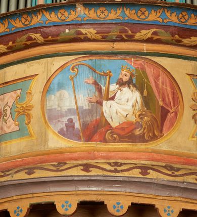 Zdjęcie nr 5: W prezbiterium w dolnej połowie ścian polichromia w formie namalowanej jasnoczerwonej, wielkowzorzytej tkaniny o motywach geometrycznych i roślinnych z niebieskim monogramem Marii i żółtymi koronami; jej górny brzeg odwiedziony jest pasem z brązowo-czerwoną dekoracją roślinną. Ściana przedzielona w połowie wysokości pasem z arkadkami wypełnionymi żółtymi wazonami oraz dekoracją roślinną na zielonym tle. W ich przyłuczach czerwone pola wypełnione malowaną roślinnością. W górnej połowie ścian malowane płyciny o jasnobrązowych ramach, których pola wypełnione są wzorem o motywie żółtych sześcioboków z fioletowymi kwiatami. Na południowej ścianie prezbiterium okna obwiedzione malowanymi, mazerowanymi ramami. W górnej części ściany, pod belkowaniem fryz utworzony z uskrzydlonych główek anielskich ukazanych na niebieskim tle, pomiędzy którymi znajdują się czerwone kampanule, pod nim podwieszone kwiatowe girlandy. Ściany zakończone profilowanym gzymsem, pomalowanym w dwa pasy – czerwony i niebieski, dekorowane motywem liści. Ściany podzielone lizenami w kolorze fioletowym, zdobionymi motywami geometrycznymi, różnymi w dolnej i górnej połowie. Strop prezbiterium z namalowaną płyciną w formie stojącego prostokąta, zamkniętego u dołu trójbocznie, o wklęsłych sześciu narożach, w których namalowana jest dekoracja roślinna. Płycina ujęta brązowo-żółtą ramą zdobioną motywem roślinnym oraz niebieskim pasem, z abstrakcyjnym motywem na wklęśnięciach. Różowe pole wypełnione białą kratką z kwiatami. W jego centrum owalny medalion w malowanej, profilowanej ramie oraz niebieskiej opasce z wypustkami, na których zawieszonych jest wokół osiem kwiatowych girland oraz cztery fantazyjne motywy kwiatowe. W medalionie namalowana scena Koronacji Matki Bożej. W jego centrum siedząca na obłokach Maria, ukazana frontalnie, z prawą ręką złożoną na piersi, lewą wyciągniętą w bok. Twarz ma pociągłą, o migdałowatych oczach, prostym nosie i pełnych ustach, okoloną długimi, falowanymi, jasnymi włosami. Ubrana jest w białą tunikę i niebieski płaszcz zarzucony na ramiona i kolana. Po jej prawej siedzi Chrystus, zwrócony w trzech czwartych w lewo, pochylony, prawą ręką podtrzymuje nad jej głową koronę. Twarz ma podłużną, o okrągłych oczach, lekko garbatym nosie i pełnych ustach, okolona brązowymi włosami i brodą. Ubrany jest w białą tunikę i czerwony płaszcz, o jego lewe ramię oparty jest krzyż. Po lewej stronie Marii siedzi Bóg Ojciec, zwrócony w trzech czwartych w prawo, lewą ręką przytrzymuje przy boku kulę, prawą podtrzymuje koronę nad głową Marii. Bóg ukazany jako starzec, o wysokim czole, podłużnej twarzy, małych oczach, garbatym nosie i pełnych ustach. Włosy i brodę ma długie i siwe. Ubrany jest w fioletową tunikę i jasnobrązowy płaszcz. Nad postaciami unosi się gołębica Ducha Świętego w glorii promienistej. Scena ukazana na tle błękitnego nieba, wypełnionego obłokami, w dolnej części obrazu z ukazanymi uskrzydlonymi główkami anielskimi. 
Ściany nawy w jednej trzeciej wysokości obwiedzione są pasem z malowanymi czerwono-brązowymi płycinami o naprzemiennym układzie owali i rombów, wypełnionymi dekoracją geometryczną i motywami roślinnymi. Ponad nimi wzdłuż ściany malowany niebieski pas o kolorowej i fantazyjnej dekoracji. Ściany w dwóch trzecich wysokości pokryte jasnoróżową polichromią o wielobarwnym, fantazyjnym, geometryczno-roślinnym wzorze z motywem równoramiennych krzyży. Ściany zakończone niebieskim fryzem przypominającym lambrekin z motywami geometrycznymi. Całość opasana profilowanym gzymsem, pomalowanym w dwa pasy – czerwony i niebieski, z dekoracją o motywach liści. Ściany podzielone lizenami pomalowanymi na kolor fioletowy, z dekoracją geometryczną. Na ścianach północnej i południowej po dwa okna obwiedzione malowanymi, mazerowanymi ramami. Pomiędzy oknami sceny w formie stojącego prostokąta zamkniętego łukiem półkolistym, w malowanej ramie dekorowanej cekinami. U dołu ramy fragment gzymsu dekorowanego kimationem z podwieszeniem zdobionym uskrzydloną główką anielską i meandrem. Rama zamknięta gzymsem wyłamanym na kształt łuku odcinkowego, podtrzymywanym na konsolach, dekorowanych motywem roślinnym. 
Na ścianie północnej scena z przedstawieniem św. Jakuba Większego z aniołem. W dolnej części obrazu święty w pozycji półleżącej, oparty o schody, ukazany frontalnie, z uniesioną głową, skierowaną w lewo i skrzyżowanymi na piersi rekami. Twarz ma owalną, o dużych oczach skierowanych w górę, prostym nosie i rozchylonych ustach, okoloną, krótkimi, jasnobrązowymi włosami i brodą. Wokół jego głowy nimb. Ubrany jest w ciemnobrązową tunikę, przepasaną sznurem, na lewym ramieniu ma muszlę, lewą ręką przytrzymuje laskę z bukłakiem. Na stopach ma sandały. Nad świętym unosi się anioł, zwrócony w trzech czwartych w lewo, w wykroku, z pochyloną głową, w prawej ręce trzyma liść palmy, w lewej, nad głową świętego - wieniec z róż. Anioł o młodzieńczej twarzy, z dużymi oczami zwróconymi w dół, dużym nosem i małymi ustami, okolonej jasnymi włosami sięgającymi ramion. Ubrany jest w żółtą tunikę oraz jasnofioletowy płaszcz. Ma białe i rozłożone skrzydła. Ukazany jest w otoczeniu obłoków. Poniżej, obok świętego na ziemi leży puklerz i miecz. Postacie ukazane we wnętrzu, z widoczną murowaną ścianą z oknami, fragmentem kolumny i schodami. W prawym dolnym sygnatura „M. Szczurowsk[i]”. 
Na ścianie południowej scena z przedstawieniem św. Izydora Oracza modlącego się przed kapliczką. W dolnej części obrazu święty w pozycji klęczącej, zwrócony profilem w prawo, z uniesionymi rękami o splecionych dłoniach w geście modlitwy, z uniesioną głową. Twarz ma szczupłą, o oczach skierowanych w górę, małym nosie i pełnych ustach, okoloną krótkimi, brązowymi włosami. Ubrany jest w białą koszulę, niebieski kaftan z czerwoną podszewką, przepasany w talii, z zawieszonym kluczem, na stopach ma buty. Przed nim, na ziemi leżą słomkowy kapelusz i bat. Przed świętym kapliczka w formie kamiennego krucyfiksu na cokole, pośród krzaków z kwiatami. Święty ukazany na tle pejzażu, z widocznym w oddali polem, na którym dwa woły ciągną pług, prowadzony przez anioła. Powyżej błękitne niebo zasnute ciemnymi chmurami, spod których wydobywa się strumień światła. W prawym dolnym rogu sygnatura „M. Szczurowski”. 
W zachodniej części nawy beżowa balustrada chóru muzycznego, na której namalowane są prostokątne płyciny, o krótszych bokach dekorowanych motywem roślinnym. Ich pola wypełnia niebieska opaska oraz różowa kratka ze zdobieniem roślinnym. Płycina środkowa w formie owalu z przedstawieniem Dawida, siedzącego na tronie i grającego na harfie. Postać ukazana do połowy, zwrócona w trzech czwartych w prawo, dwiema wyciągniętymi rękami dotyka strun harfy. Dawid o pociągłej twarzy, ze schematycznie oddanymi rysami, okolony brązowymi włosami i brodą. Na głowie ma koronę otwartą. Ubrany jest w czerwoną suknię, na ramiona ma narzuconą gronostajową pelerynę. Nad nim zawieszony czerwony baldachim, z zieloną podszewką. W oddali widoczny zarys zabudowań miejskich. Profile cokołu i gzymsu balustrady malowane w kolorowe pasy, z dekoracją roślinną. 
Na stropie nawy malowana płycina w formie stojącego prostokąta o zaokrąglonych narożach, ujęta niebieską ramą o geometrycznym wzorze, w miejscu naroży stylizowane roślinne palmety. W narożach płyciny cztery owalne medaliony z popiersiami św. Stanisława Kostki, św. Kazimierza Jagiellończyka, św. Stanisława z Piotrowinem i św. Jana Kantego. Medaliony ujęte są wolutowymi kartuszami, dekorowanymi stylizowanym ornamentem roślinnym. W polu centralnym malowanej płyciny scena ze św. Klemensem w formie stojącego prostokąta o wklęsłych narożach, ujęta malowaną ramą, w narożach uskrzydlone główki anielskie. Scena otoczona drugą ramą o fantazyjnym zarysie, dekorowaną na bokach fantazyjnymi szczytami z ornamentem roślinnym, które połączone są kwiecistymi girlandami z narożnymi kartuszami. Scena ze św. Klemensem przedstawia go w kamieniołomie, błogosławiącego robotników z kilofami. Święty ukazany jest jako starzec, z siwą brodą i włosami oraz łysiną na głowie. Ubrany jest w jasnofioletową sutannę, białą rokietę z dekoracyjnym obszyciem oraz jasnoczerwony, szeroki ornat, na piersi ma paliusz. W tle pomiędzy dwoma wzniesieniami skalnymi widoczne morze, z płynącym statkiem, w oddali zaś brzeg z domem. Na skarpie w lewej części obrazu ukazany baranek w glorii promienistej, na przeciwległej dwaj żołnierze, którzy zrzucili świętego z kotwicą zawieszoną u szyi do morza. Przy dolnej krawędzi obrazu sygnatura „pinxit 1910 / M. Szczurowski”.
