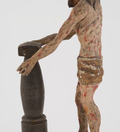 Zdjęcie nr 6: Pełnoplastyczna figurka Chrystusa z ruchomymi rękami, stojącego przy słupie ustawiona jest na owalnej podstawie. Chrystus stoi w wykroku, delikatnie pochylony ku słupowi, z głową skierowaną w prawo, z rękami wysuniętymi przed siebie, ujmującymi słup biczowania. Twarz ma szczupłą, pociągłą, o uwypuklonych kościach policzkowych, dużych oczach i nosie, okoloną krótką brodą i długimi, falowanymi włosami opadającymi na plecy. Ciało Chrystusa jest wychudzone, z silnie uwypuklonymi żebrami klatki piersiowej. Przez biodra ma przewiązane krótkie, obficie drapowane, złocone perizonium ze zwisem tkaniny na prawym boku. Słup w formie tralki na cokole. Polichromia ciała naturalistyczna, z zaznaczonymi licznymi strużkami krwi. 
