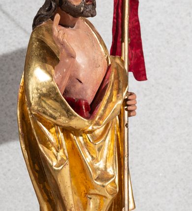 Zdjęcie nr 2: Figura pełnoplastyczna, ustawiona na rzeźbionym fragmencie podłoża i na niskiej podstawie na planie ośmioboku. Chrystus zwrócony jest frontalnie, z głową skierowaną w prawo. W lewej ręce trzyma drzewce zakończone krzyżem z chorągwią z tkaniny z naszytym krzyżem, prawą rękę unosi w geście błogosławieństwa. Twarz ma owalną, o dużych, półprzymkniętych oczach, uniesionych brwiach, dużym nosie i małych ustach, okoloną krótką brodą skręconą w dwa pukle oraz brązowymi włosami sięgającymi ramion, rzeźbionymi w regularne fale. Chrystus ubrany jest w złocony płaszcz z czerwoną podszewką zarzucony na ramiona i plecy, otaczający postać i zarzucony z przodu na lewe przedramię, odsłaniając nagi tors z zaznaczoną raną w boku. Polichromia ciała naturalistyczna.
