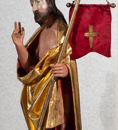 Zdjęcie nr 3: Figura pełnoplastyczna, ustawiona na rzeźbionym fragmencie podłoża i na niskiej podstawie na planie ośmioboku. Chrystus zwrócony jest frontalnie, z głową skierowaną w prawo. W lewej ręce trzyma drzewce zakończone krzyżem z chorągwią z tkaniny z naszytym krzyżem, prawą rękę unosi w geście błogosławieństwa. Twarz ma owalną, o dużych, półprzymkniętych oczach, uniesionych brwiach, dużym nosie i małych ustach, okoloną krótką brodą skręconą w dwa pukle oraz brązowymi włosami sięgającymi ramion, rzeźbionymi w regularne fale. Chrystus ubrany jest w złocony płaszcz z czerwoną podszewką zarzucony na ramiona i plecy, otaczający postać i zarzucony z przodu na lewe przedramię, odsłaniając nagi tors z zaznaczoną raną w boku. Polichromia ciała naturalistyczna.
