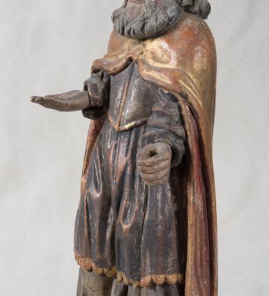 Zdjęcie nr 5: Rzeźba pełnoplastyczna, nieopracowana z tyłu, ustawiona na podstawie o nieregularnym kształcie. Święty zwrócony jest frontalnie, w delikatnym kontrapoście, z obiema rękami ugiętymi i wyciągniętymi do przodu. Prawa dłoń wysunięta zwrócona ku górze, w lewej trzyma laskę zakończoną krzyżem na kuli. Twarz ma kwadratową, o dużych oczach, krótkim nosie i małych, pełnych ustach, okoloną gęstą siwą brodą oraz falowanymi włosami, sięgającymi ramion. Mikołaj ubrany jest w długą, zieloną albę, niebieską rokietę zdobioną u dołu oraz zarzucony na ramiona, spięty na piersiach klamrą, pozłacany płaszcz, na głowie ma złoconą infułę. Polichromia ciała naturalistyczna, o jasnej, lekko zaróżowionej karnacji.
