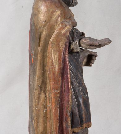 Zdjęcie nr 9: Rzeźba pełnoplastyczna, nieopracowana z tyłu, ustawiona na podstawie o nieregularnym kształcie. Święty zwrócony jest frontalnie, w delikatnym kontrapoście, z obiema rękami ugiętymi i wyciągniętymi do przodu. Prawa dłoń wysunięta zwrócona ku górze, w lewej trzyma laskę zakończoną krzyżem na kuli. Twarz ma kwadratową, o dużych oczach, krótkim nosie i małych, pełnych ustach, okoloną gęstą siwą brodą oraz falowanymi włosami, sięgającymi ramion. Mikołaj ubrany jest w długą, zieloną albę, niebieską rokietę zdobioną u dołu oraz zarzucony na ramiona, spięty na piersiach klamrą, pozłacany płaszcz, na głowie ma złoconą infułę. Polichromia ciała naturalistyczna, o jasnej, lekko zaróżowionej karnacji.
