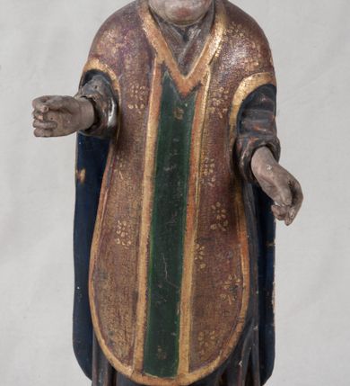 Zdjęcie nr 4: Rzeźba pełnoplastyczna, nieopracowana z tyłu, ustawiona jest na zielonej podstawie o nieregularnym kształcie. Święty zwrócony jest frontalnie, z obiema rękami ugiętymi, wyciągniętymi do przodu. Ma kwadratową twarz, o podkreślonej linii żuchwy i brody, o dużych oczach, krótkim nosie i małych, pełnych ustach. Włosy ma brązowe, falowane sięgające ramion. Ubrany jest w szarą, silnie drapowaną u dołu albę, widoczny u szyi humerał oraz złocony ornat skrzypcowy o wzorze kwiatowym, z pretekstą przednią w kolorze zielonym i niebieską podszewką. Na głowie ma złoconą mitrę. Polichromia ciała naturalistyczna o jasnej, zaróżowionej karnacji w partii twarzy.