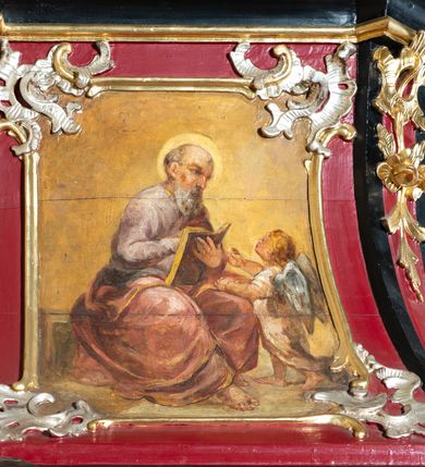 Zdjęcie nr 4: Ambona przyścienna, wisząca z dostępem od zakrystii. Podwieszenie w formie odwróconego ostrosłupa o ściętym wierzchołku, na krawędziach flankowane pilastrami zakończonymi u góry wolutowo, a na ściankach zdobione płycinami. Kosz na planie kwadratu umieszczony na wybrzuszonej podstawie, dekorowanej rocaille'owymi kartuszami, o ściętych górnych narożach; ścianki wklęsłe, zwężające się ku górze, ujęte w narożach pilastrami z trzonami dekorowanymi rocaille'em i zwisami z różanych gałązek. Podpory dźwigają gzyms wyłamany na ich osi, tworzący parapet. W polu ścianek płyciny z rocaille'ami w narożach z malowanymi olejem na desce obrazami przedstawiającymi ewangelistów. Od strony lewej: św. Mateusz z aniołem, pośrodku św. Marek z lwem i św. Łukasz z wołem, a po prawej św. Jan z orłem. Ewangeliści ukazani w pozycji siedzącej z księgami w dłoniach, ubrani w różnokolorowe, powłóczyste szaty na złotym tle. Zaplecek w kształcie stojącego prostokąta, ujęty pilastrami dekorowanymi rocaille'ami. W polu płycina w kształcie stojącego prostokąta, zamkniętego łukiem nadwieszonym z płaskorzeźbionymi tablicami dekalogu, ujętymi od dołu skrzyżowanymi gałązkami oliwnymi, związanymi wstęgą. Belkowanie nad zapleckiem o wypukłej linii, z przerwanym pośrodku gzymsem, od góry wypełnionym rocaille'em, od dołu ozdobionym podwieszonym lambrekinem z chwostami. Zwieńczenie ma formę zbliżoną do trapezu, ujęte jest wolutami spływającymi na belkowanie, na których ustawione są rokokowe wazony. W polu zwieńczenia znajduje się medalion z glorią pośród obłoków, a powyżej gołębica Ducha Świętego. Zwieńczenie flankują umocowane na ścianie, uskrzydlone główki aniołków dobrej i złej rady: po lewej o jasnej karnacji, a po prawej o czarnej karnacji. Szczyt zwieńczenia ma formę gzymsu z podwieszonym lambrekinem. Całość wieńczy zawieszona na ścianie promienista gloria z wieńcem utworzonym z obłoków. Struktura polichromowana jest w kolorze czerwonym, z czarnymi profilami oraz złoconą i srebrzoną ornamentyką. 
