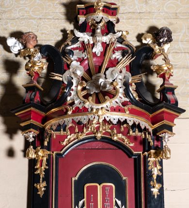 Zdjęcie nr 7: Ambona przyścienna, wisząca z dostępem od zakrystii. Podwieszenie w formie odwróconego ostrosłupa o ściętym wierzchołku, na krawędziach flankowane pilastrami zakończonymi u góry wolutowo, a na ściankach zdobione płycinami. Kosz na planie kwadratu umieszczony na wybrzuszonej podstawie, dekorowanej rocaille'owymi kartuszami, o ściętych górnych narożach; ścianki wklęsłe, zwężające się ku górze, ujęte w narożach pilastrami z trzonami dekorowanymi rocaille'em i zwisami z różanych gałązek. Podpory dźwigają gzyms wyłamany na ich osi, tworzący parapet. W polu ścianek płyciny z rocaille'ami w narożach z malowanymi olejem na desce obrazami przedstawiającymi ewangelistów. Od strony lewej: św. Mateusz z aniołem, pośrodku św. Marek z lwem i św. Łukasz z wołem, a po prawej św. Jan z orłem. Ewangeliści ukazani w pozycji siedzącej z księgami w dłoniach, ubrani w różnokolorowe, powłóczyste szaty na złotym tle. Zaplecek w kształcie stojącego prostokąta, ujęty pilastrami dekorowanymi rocaille'ami. W polu płycina w kształcie stojącego prostokąta, zamkniętego łukiem nadwieszonym z płaskorzeźbionymi tablicami dekalogu, ujętymi od dołu skrzyżowanymi gałązkami oliwnymi, związanymi wstęgą. Belkowanie nad zapleckiem o wypukłej linii, z przerwanym pośrodku gzymsem, od góry wypełnionym rocaille'em, od dołu ozdobionym podwieszonym lambrekinem z chwostami. Zwieńczenie ma formę zbliżoną do trapezu, ujęte jest wolutami spływającymi na belkowanie, na których ustawione są rokokowe wazony. W polu zwieńczenia znajduje się medalion z glorią pośród obłoków, a powyżej gołębica Ducha Świętego. Zwieńczenie flankują umocowane na ścianie, uskrzydlone główki aniołków dobrej i złej rady: po lewej o jasnej karnacji, a po prawej o czarnej karnacji. Szczyt zwieńczenia ma formę gzymsu z podwieszonym lambrekinem. Całość wieńczy zawieszona na ścianie promienista gloria z wieńcem utworzonym z obłoków. Struktura polichromowana jest w kolorze czerwonym, z czarnymi profilami oraz złoconą i srebrzoną ornamentyką. 
