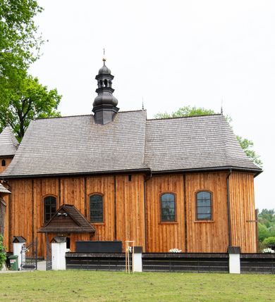 Zdjęcie nr 3: Kościół orientowany, drewniany o konstrukcji zrębowej z wieżą o konstrukcji słupowo-ryglowej na rzucie kwadratu. Nawa prostokątna z węższym, prostokątnym prezbiterium zamkniętym trójboczną absydą. Przy prezbiterium, od północy znajduje się zakrystia, a przy nawie, od południa – kruchta. Na osi nawy, od zachodu wieża o pochyłych ścianach z nadwieszoną izbicą. W kościele sześć otworów okiennych w kształcie stojących prostokątów, ujętych opaskami z prostych listew: dwa od strony północnej i cztery od strony południowej. Na zewnątrz okien zamontowane są współczesne kraty z ozdobnych prętów. Główne wejście do kościoła od zachodu w przyziemiu wieży. Dach dwuspadowy, od wschodu trójspadowy, wykonany w konstrukcji krokwiowo-jętkowej z podwójnymi jętkami, kryty gontem. Zakrystia zadaszona przedłużoną połacią głównego dachu. Na osi kalenicy dachu wieżyczka na sygnaturkę, barokowa z latarnią, kryta blachą miedzianą. Z zewnątrz ściany kościoła i wieży pokryto szalunkiem pionowym z listwami na stykach łączenia desek. Nadwieszona izbica wieży z wyciętymi ornamentami na dolnych zakończeniach desek. W czterech ścianach izbicy po dwa okna w kształcie stojącego prostokąta, zamknięte łukiem nadwieszonym. Wieża nakryta daszkiem czterospadowym, krytym gontem. Do obu stron wieży dostawione nowe dobudówki: w południowej umieszczono schody na chór muzyczny, a w północnej znajdują się pomieszczenia gospodarcze. Kościół w przyziemiu jest podbudowany betonowym cokołem, silnie wysuniętym przed lico ścian i zabezpieczony małym daszkiem.

