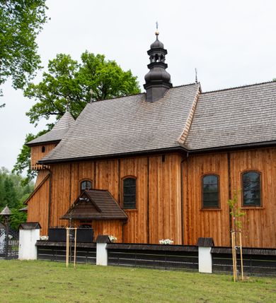 Zdjęcie nr 4: Kościół orientowany, drewniany o konstrukcji zrębowej z wieżą o konstrukcji słupowo-ryglowej na rzucie kwadratu. Nawa prostokątna z węższym, prostokątnym prezbiterium zamkniętym trójboczną absydą. Przy prezbiterium, od północy znajduje się zakrystia, a przy nawie, od południa – kruchta. Na osi nawy, od zachodu wieża o pochyłych ścianach z nadwieszoną izbicą. W kościele sześć otworów okiennych w kształcie stojących prostokątów, ujętych opaskami z prostych listew: dwa od strony północnej i cztery od strony południowej. Na zewnątrz okien zamontowane są współczesne kraty z ozdobnych prętów. Główne wejście do kościoła od zachodu w przyziemiu wieży. Dach dwuspadowy, od wschodu trójspadowy, wykonany w konstrukcji krokwiowo-jętkowej z podwójnymi jętkami, kryty gontem. Zakrystia zadaszona przedłużoną połacią głównego dachu. Na osi kalenicy dachu wieżyczka na sygnaturkę, barokowa z latarnią, kryta blachą miedzianą. Z zewnątrz ściany kościoła i wieży pokryto szalunkiem pionowym z listwami na stykach łączenia desek. Nadwieszona izbica wieży z wyciętymi ornamentami na dolnych zakończeniach desek. W czterech ścianach izbicy po dwa okna w kształcie stojącego prostokąta, zamknięte łukiem nadwieszonym. Wieża nakryta daszkiem czterospadowym, krytym gontem. Do obu stron wieży dostawione nowe dobudówki: w południowej umieszczono schody na chór muzyczny, a w północnej znajdują się pomieszczenia gospodarcze. Kościół w przyziemiu jest podbudowany betonowym cokołem, silnie wysuniętym przed lico ścian i zabezpieczony małym daszkiem.

