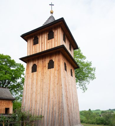 Zdjęcie nr 1: Dzwonnica na planie kwadratu, podpiwniczona, dwukondygnacyjna, drewniana, oszalowana deskami z listwowaniem. Kondygnacja dolna na niskim cokole, o ścianach zwężających się ku górze, z wejściem od zachodu, na trzech ścianach przepruta otworami okiennymi (dwoma na każdej ścianie), nakryta pulpitowym daszkiem. Kondygnacja górna o ścianach prostych, z dwoma małymi otworami okiennymi na każdej ścianie. Otwory okienne w kształcie stojących prostokątów, zamkniętych łukiem nadwieszonym. Dzwonnica nakryta dachem półszczytowym zakończonym złoconą kulą i zatkniętym na niej łacińskim krzyżem.