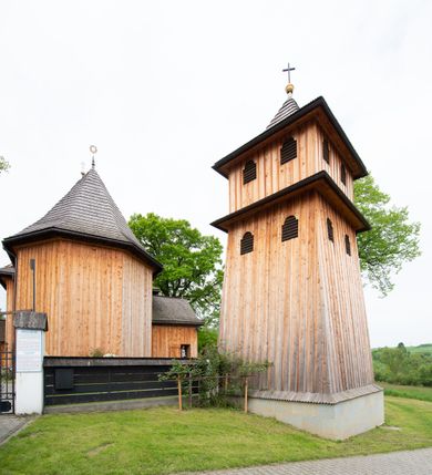Zdjęcie nr 2: Dzwonnica na planie kwadratu, podpiwniczona, dwukondygnacyjna, drewniana, oszalowana deskami z listwowaniem. Kondygnacja dolna na niskim cokole, o ścianach zwężających się ku górze, z wejściem od zachodu, na trzech ścianach przepruta otworami okiennymi (dwoma na każdej ścianie), nakryta pulpitowym daszkiem. Kondygnacja górna o ścianach prostych, z dwoma małymi otworami okiennymi na każdej ścianie. Otwory okienne w kształcie stojących prostokątów, zamkniętych łukiem nadwieszonym. Dzwonnica nakryta dachem półszczytowym zakończonym złoconą kulą i zatkniętym na niej łacińskim krzyżem.