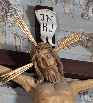 Zdjęcie nr 5: Figura Chrystusa przybita do krzyża trzema gwoździami pełna, niedrążona. Ciało w niewielkim zwisie, głowa delikatnie opada na prawy bark, stopy skrzyżowane w układzie prawa na lewą. Sylwetka podkreślona wydatną klatką piersiową, z wyraźnie zaznaczoną linią żeber i mostka, a także z precyzyjnie ukazaną anatomią. Twarz podłużna, z długim nosem, przymkniętymi oczami, okolona brodą ułożoną w długie, równolegle opadające, proste pukle. Włosy długie, brązowe, spływające na plecy oraz z przodu dwoma, silnie skręconymi lokami. Na głowie zielona korona cierniowa, a wokół niej nimb krzyżowy złożony z trzech wiązek promieni. Perizonium ciasno oplatające biodra, z dwoma zwisami po bokach, po lewej stronie dłuższym. Polichromia ciała naturalistyczna z delikatnie zaznaczonymi śladami męki, perizonium, gwoździe i nimb złocone. Nad głową Chrystusa titulus w formie pionowej banderoli z napisem „J[esus] N[azarenus] / R[ex] J[udaeorum]”.