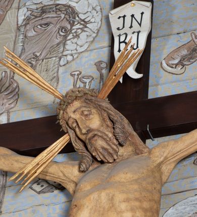 Zdjęcie nr 2: Figura Chrystusa przybita do krzyża trzema gwoździami pełna, niedrążona. Ciało w niewielkim zwisie, głowa delikatnie opada na prawy bark, stopy skrzyżowane w układzie prawa na lewą. Sylwetka podkreślona wydatną klatką piersiową, z wyraźnie zaznaczoną linią żeber i mostka, a także z precyzyjnie ukazaną anatomią. Twarz podłużna, z długim nosem, przymkniętymi oczami, okolona brodą ułożoną w długie, równolegle opadające, proste pukle. Włosy długie, brązowe, spływające na plecy oraz z przodu dwoma, silnie skręconymi lokami. Na głowie zielona korona cierniowa, a wokół niej nimb krzyżowy złożony z trzech wiązek promieni. Perizonium ciasno oplatające biodra, z dwoma zwisami po bokach, po lewej stronie dłuższym. Polichromia ciała naturalistyczna z delikatnie zaznaczonymi śladami męki, perizonium, gwoździe i nimb złocone. Nad głową Chrystusa titulus w formie pionowej banderoli z napisem „J[esus] N[azarenus] / R[ex] J[udaeorum]”.