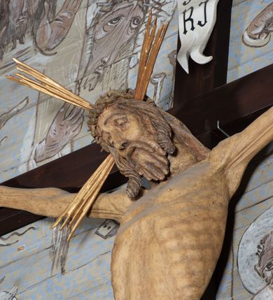 Zdjęcie nr 8: Figura Chrystusa przybita do krzyża trzema gwoździami pełna, niedrążona. Ciało w niewielkim zwisie, głowa delikatnie opada na prawy bark, stopy skrzyżowane w układzie prawa na lewą. Sylwetka podkreślona wydatną klatką piersiową, z wyraźnie zaznaczoną linią żeber i mostka, a także z precyzyjnie ukazaną anatomią. Twarz podłużna, z długim nosem, przymkniętymi oczami, okolona brodą ułożoną w długie, równolegle opadające, proste pukle. Włosy długie, brązowe, spływające na plecy oraz z przodu dwoma, silnie skręconymi lokami. Na głowie zielona korona cierniowa, a wokół niej nimb krzyżowy złożony z trzech wiązek promieni. Perizonium ciasno oplatające biodra, z dwoma zwisami po bokach, po lewej stronie dłuższym. Polichromia ciała naturalistyczna z delikatnie zaznaczonymi śladami męki, perizonium, gwoździe i nimb złocone. Nad głową Chrystusa titulus w formie pionowej banderoli z napisem „J[esus] N[azarenus] / R[ex] J[udaeorum]”.