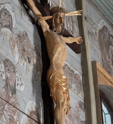 Zdjęcie nr 9: Figura Chrystusa przybita do krzyża trzema gwoździami pełna, niedrążona. Ciało w niewielkim zwisie, głowa delikatnie opada na prawy bark, stopy skrzyżowane w układzie prawa na lewą. Sylwetka podkreślona wydatną klatką piersiową, z wyraźnie zaznaczoną linią żeber i mostka, a także z precyzyjnie ukazaną anatomią. Twarz podłużna, z długim nosem, przymkniętymi oczami, okolona brodą ułożoną w długie, równolegle opadające, proste pukle. Włosy długie, brązowe, spływające na plecy oraz z przodu dwoma, silnie skręconymi lokami. Na głowie zielona korona cierniowa, a wokół niej nimb krzyżowy złożony z trzech wiązek promieni. Perizonium ciasno oplatające biodra, z dwoma zwisami po bokach, po lewej stronie dłuższym. Polichromia ciała naturalistyczna z delikatnie zaznaczonymi śladami męki, perizonium, gwoździe i nimb złocone. Nad głową Chrystusa titulus w formie pionowej banderoli z napisem „J[esus] N[azarenus] / R[ex] J[udaeorum]”.