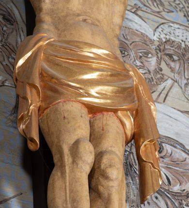 Zdjęcie nr 10: Figura Chrystusa przybita do krzyża trzema gwoździami pełna, niedrążona. Ciało w niewielkim zwisie, głowa delikatnie opada na prawy bark, stopy skrzyżowane w układzie prawa na lewą. Sylwetka podkreślona wydatną klatką piersiową, z wyraźnie zaznaczoną linią żeber i mostka, a także z precyzyjnie ukazaną anatomią. Twarz podłużna, z długim nosem, przymkniętymi oczami, okolona brodą ułożoną w długie, równolegle opadające, proste pukle. Włosy długie, brązowe, spływające na plecy oraz z przodu dwoma, silnie skręconymi lokami. Na głowie zielona korona cierniowa, a wokół niej nimb krzyżowy złożony z trzech wiązek promieni. Perizonium ciasno oplatające biodra, z dwoma zwisami po bokach, po lewej stronie dłuższym. Polichromia ciała naturalistyczna z delikatnie zaznaczonymi śladami męki, perizonium, gwoździe i nimb złocone. Nad głową Chrystusa titulus w formie pionowej banderoli z napisem „J[esus] N[azarenus] / R[ex] J[udaeorum]”.