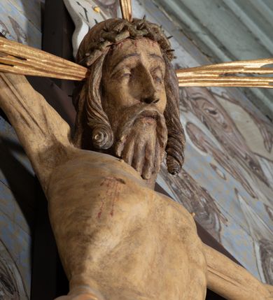 Zdjęcie nr 12: Figura Chrystusa przybita do krzyża trzema gwoździami pełna, niedrążona. Ciało w niewielkim zwisie, głowa delikatnie opada na prawy bark, stopy skrzyżowane w układzie prawa na lewą. Sylwetka podkreślona wydatną klatką piersiową, z wyraźnie zaznaczoną linią żeber i mostka, a także z precyzyjnie ukazaną anatomią. Twarz podłużna, z długim nosem, przymkniętymi oczami, okolona brodą ułożoną w długie, równolegle opadające, proste pukle. Włosy długie, brązowe, spływające na plecy oraz z przodu dwoma, silnie skręconymi lokami. Na głowie zielona korona cierniowa, a wokół niej nimb krzyżowy złożony z trzech wiązek promieni. Perizonium ciasno oplatające biodra, z dwoma zwisami po bokach, po lewej stronie dłuższym. Polichromia ciała naturalistyczna z delikatnie zaznaczonymi śladami męki, perizonium, gwoździe i nimb złocone. Nad głową Chrystusa titulus w formie pionowej banderoli z napisem „J[esus] N[azarenus] / R[ex] J[udaeorum]”.