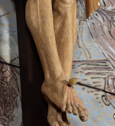 Zdjęcie nr 13: Figura Chrystusa przybita do krzyża trzema gwoździami pełna, niedrążona. Ciało w niewielkim zwisie, głowa delikatnie opada na prawy bark, stopy skrzyżowane w układzie prawa na lewą. Sylwetka podkreślona wydatną klatką piersiową, z wyraźnie zaznaczoną linią żeber i mostka, a także z precyzyjnie ukazaną anatomią. Twarz podłużna, z długim nosem, przymkniętymi oczami, okolona brodą ułożoną w długie, równolegle opadające, proste pukle. Włosy długie, brązowe, spływające na plecy oraz z przodu dwoma, silnie skręconymi lokami. Na głowie zielona korona cierniowa, a wokół niej nimb krzyżowy złożony z trzech wiązek promieni. Perizonium ciasno oplatające biodra, z dwoma zwisami po bokach, po lewej stronie dłuższym. Polichromia ciała naturalistyczna z delikatnie zaznaczonymi śladami męki, perizonium, gwoździe i nimb złocone. Nad głową Chrystusa titulus w formie pionowej banderoli z napisem „J[esus] N[azarenus] / R[ex] J[udaeorum]”.