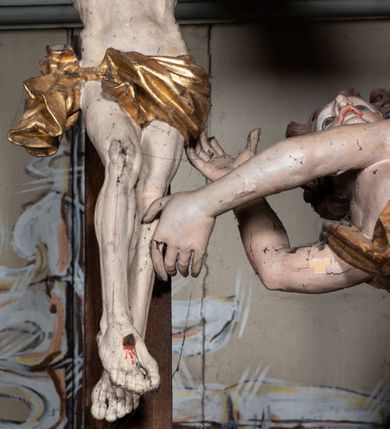Zdjęcie nr 7: Rzeźbiarska grupa Ukrzyżowania, przedstawiająca przybitego do krzyża Chrystusa podtrzymywanego przez trzy anioły. Figura Chrystusa ścięta z tyłu, przybita do krzyża trzema gwoździami, głowa opadła na prawe ramię, ramiona szeroko rozłożone w delikatnym zwisie. Ciało szczupłe, o wyraźnie podkreślonej anatomii, wydatna klata piersiowa z delikatnie zaznaczoną linią żeber. Twarz o sumarycznie opracowanych rysach, z krótkim, ciemnobrązowym zarostem, dużymi ustami i zamkniętymi oczami. Włosy długie, ciemnobrązowe, falowane, opadające na plecy i prawe ramię grubymi lokami. Na głowie złocona korona cierniowa. Na biodrach krótkie, złocone, przewiązane sznurem perizonium z węzłem i krótkim zwisem na prawym boku. Na czole, dłoniach, stopach i prawym boku delikatne strużki krwi. Kolorystyka ciała naturalistyczna. Krzyż polichromowany na ciemny brąz, nad głową Chrystusa złocony titulus w formie podłużnej banderoli z napisem „IN / RI”. Po prawej stronie, w połowie wysokości dolnej części pionowej belki pełnoplastyczna figura anioła, unoszącego się w powietrzu, z rękami zwróconymi do podtrzymywania krucyfiksu. Głowa w trzech czwartych skierowana w lewo, odchylona do tyłu. Twarz owalna, o wyrazistych rysach, z mocno zaróżowionymi policzkami. Włosy krótkie, kręcone i brązowe. Ubrany jest w złoconą szatę odsłaniającą ramiona, fragment klatki piersiowej i nogi do wysokości kolan. Na plecach ma duże, złocone skrzydła. Belka pozioma flankowana parą przeciwstawnych, unoszących się w powietrzu, pełnoplastycznych putt, podtrzymujących dłońmi krawędzie poziomej belki krzyża. Główki zwrócone frontalnie, twarze o zaokrąglonych, pełnych, wyrazistych rysach i zarumienionych policzkach z dużymi oczami. Włosy krótkie, kręcone i brązowe. Ciała nagie, o zaokrąglonych kształtach, z rozwianymi na plecach, niewielkimi, złoconymi płaszczami i parami małych skrzydełek. Polichromia w odsłoniętych partiach ciała naturalistyczna, szaty i detale złocone.
