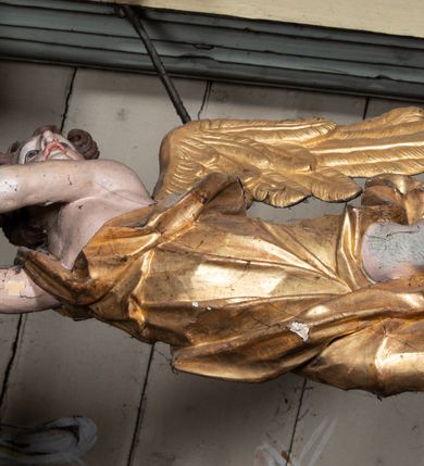 Zdjęcie nr 6: Rzeźbiarska grupa Ukrzyżowania, przedstawiająca przybitego do krzyża Chrystusa podtrzymywanego przez trzy anioły. Figura Chrystusa ścięta z tyłu, przybita do krzyża trzema gwoździami, głowa opadła na prawe ramię, ramiona szeroko rozłożone w delikatnym zwisie. Ciało szczupłe, o wyraźnie podkreślonej anatomii, wydatna klata piersiowa z delikatnie zaznaczoną linią żeber. Twarz o sumarycznie opracowanych rysach, z krótkim, ciemnobrązowym zarostem, dużymi ustami i zamkniętymi oczami. Włosy długie, ciemnobrązowe, falowane, opadające na plecy i prawe ramię grubymi lokami. Na głowie złocona korona cierniowa. Na biodrach krótkie, złocone, przewiązane sznurem perizonium z węzłem i krótkim zwisem na prawym boku. Na czole, dłoniach, stopach i prawym boku delikatne strużki krwi. Kolorystyka ciała naturalistyczna. Krzyż polichromowany na ciemny brąz, nad głową Chrystusa złocony titulus w formie podłużnej banderoli z napisem „IN / RI”. Po prawej stronie, w połowie wysokości dolnej części pionowej belki pełnoplastyczna figura anioła, unoszącego się w powietrzu, z rękami zwróconymi do podtrzymywania krucyfiksu. Głowa w trzech czwartych skierowana w lewo, odchylona do tyłu. Twarz owalna, o wyrazistych rysach, z mocno zaróżowionymi policzkami. Włosy krótkie, kręcone i brązowe. Ubrany jest w złoconą szatę odsłaniającą ramiona, fragment klatki piersiowej i nogi do wysokości kolan. Na plecach ma duże, złocone skrzydła. Belka pozioma flankowana parą przeciwstawnych, unoszących się w powietrzu, pełnoplastycznych putt, podtrzymujących dłońmi krawędzie poziomej belki krzyża. Główki zwrócone frontalnie, twarze o zaokrąglonych, pełnych, wyrazistych rysach i zarumienionych policzkach z dużymi oczami. Włosy krótkie, kręcone i brązowe. Ciała nagie, o zaokrąglonych kształtach, z rozwianymi na plecach, niewielkimi, złoconymi płaszczami i parami małych skrzydełek. Polichromia w odsłoniętych partiach ciała naturalistyczna, szaty i detale złocone.