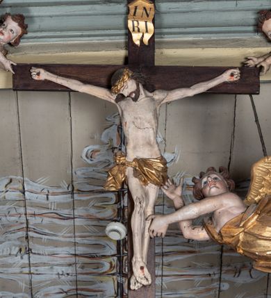 Zdjęcie nr 1: Rzeźbiarska grupa Ukrzyżowania, przedstawiająca przybitego do krzyża Chrystusa podtrzymywanego przez trzy anioły. Figura Chrystusa ścięta z tyłu, przybita do krzyża trzema gwoździami, głowa opadła na prawe ramię, ramiona szeroko rozłożone w delikatnym zwisie. Ciało szczupłe, o wyraźnie podkreślonej anatomii, wydatna klata piersiowa z delikatnie zaznaczoną linią żeber. Twarz o sumarycznie opracowanych rysach, z krótkim, ciemnobrązowym zarostem, dużymi ustami i zamkniętymi oczami. Włosy długie, ciemnobrązowe, falowane, opadające na plecy i prawe ramię grubymi lokami. Na głowie złocona korona cierniowa. Na biodrach krótkie, złocone, przewiązane sznurem perizonium z węzłem i krótkim zwisem na prawym boku. Na czole, dłoniach, stopach i prawym boku delikatne strużki krwi. Kolorystyka ciała naturalistyczna. Krzyż polichromowany na ciemny brąz, nad głową Chrystusa złocony titulus w formie podłużnej banderoli z napisem „IN / RI”. Po prawej stronie, w połowie wysokości dolnej części pionowej belki pełnoplastyczna figura anioła, unoszącego się w powietrzu, z rękami zwróconymi do podtrzymywania krucyfiksu. Głowa w trzech czwartych skierowana w lewo, odchylona do tyłu. Twarz owalna, o wyrazistych rysach, z mocno zaróżowionymi policzkami. Włosy krótkie, kręcone i brązowe. Ubrany jest w złoconą szatę odsłaniającą ramiona, fragment klatki piersiowej i nogi do wysokości kolan. Na plecach ma duże, złocone skrzydła. Belka pozioma flankowana parą przeciwstawnych, unoszących się w powietrzu, pełnoplastycznych putt, podtrzymujących dłońmi krawędzie poziomej belki krzyża. Główki zwrócone frontalnie, twarze o zaokrąglonych, pełnych, wyrazistych rysach i zarumienionych policzkach z dużymi oczami. Włosy krótkie, kręcone i brązowe. Ciała nagie, o zaokrąglonych kształtach, z rozwianymi na plecach, niewielkimi, złoconymi płaszczami i parami małych skrzydełek. Polichromia w odsłoniętych partiach ciała naturalistyczna, szaty i detale złocone.