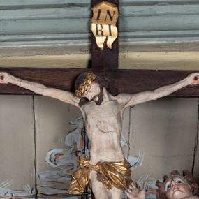 Zdjęcie nr 1: Rzeźbiarska grupa Ukrzyżowania, przedstawiająca przybitego do krzyża Chrystusa podtrzymywanego przez trzy anioły. Figura Chrystusa ścięta z tyłu, przybita do krzyża trzema gwoździami, głowa opadła na prawe ramię, ramiona szeroko rozłożone w delikatnym zwisie. Ciało szczupłe, o wyraźnie podkreślonej anatomii, wydatna klata piersiowa z delikatnie zaznaczoną linią żeber. Twarz o sumarycznie opracowanych rysach, z krótkim, ciemnobrązowym zarostem, dużymi ustami i zamkniętymi oczami. Włosy długie, ciemnobrązowe, falowane, opadające na plecy i prawe ramię grubymi lokami. Na głowie złocona korona cierniowa. Na biodrach krótkie, złocone, przewiązane sznurem perizonium z węzłem i krótkim zwisem na prawym boku. Na czole, dłoniach, stopach i prawym boku delikatne strużki krwi. Kolorystyka ciała naturalistyczna. Krzyż polichromowany na ciemny brąz, nad głową Chrystusa złocony titulus w formie podłużnej banderoli z napisem „IN / RI”. Po prawej stronie, w połowie wysokości dolnej części pionowej belki pełnoplastyczna figura anioła, unoszącego się w powietrzu, z rękami zwróconymi do podtrzymywania krucyfiksu. Głowa w trzech czwartych skierowana w lewo, odchylona do tyłu. Twarz owalna, o wyrazistych rysach, z mocno zaróżowionymi policzkami. Włosy krótkie, kręcone i brązowe. Ubrany jest w złoconą szatę odsłaniającą ramiona, fragment klatki piersiowej i nogi do wysokości kolan. Na plecach ma duże, złocone skrzydła. Belka pozioma flankowana parą przeciwstawnych, unoszących się w powietrzu, pełnoplastycznych putt, podtrzymujących dłońmi krawędzie poziomej belki krzyża. Główki zwrócone frontalnie, twarze o zaokrąglonych, pełnych, wyrazistych rysach i zarumienionych policzkach z dużymi oczami. Włosy krótkie, kręcone i brązowe. Ciała nagie, o zaokrąglonych kształtach, z rozwianymi na plecach, niewielkimi, złoconymi płaszczami i parami małych skrzydełek. Polichromia w odsłoniętych partiach ciała naturalistyczna, szaty i detale złocone.