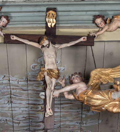 Zdjęcie nr 2: Rzeźbiarska grupa Ukrzyżowania, przedstawiająca przybitego do krzyża Chrystusa podtrzymywanego przez trzy anioły. Figura Chrystusa ścięta z tyłu, przybita do krzyża trzema gwoździami, głowa opadła na prawe ramię, ramiona szeroko rozłożone w delikatnym zwisie. Ciało szczupłe, o wyraźnie podkreślonej anatomii, wydatna klata piersiowa z delikatnie zaznaczoną linią żeber. Twarz o sumarycznie opracowanych rysach, z krótkim, ciemnobrązowym zarostem, dużymi ustami i zamkniętymi oczami. Włosy długie, ciemnobrązowe, falowane, opadające na plecy i prawe ramię grubymi lokami. Na głowie złocona korona cierniowa. Na biodrach krótkie, złocone, przewiązane sznurem perizonium z węzłem i krótkim zwisem na prawym boku. Na czole, dłoniach, stopach i prawym boku delikatne strużki krwi. Kolorystyka ciała naturalistyczna. Krzyż polichromowany na ciemny brąz, nad głową Chrystusa złocony titulus w formie podłużnej banderoli z napisem „IN / RI”. Po prawej stronie, w połowie wysokości dolnej części pionowej belki pełnoplastyczna figura anioła, unoszącego się w powietrzu, z rękami zwróconymi do podtrzymywania krucyfiksu. Głowa w trzech czwartych skierowana w lewo, odchylona do tyłu. Twarz owalna, o wyrazistych rysach, z mocno zaróżowionymi policzkami. Włosy krótkie, kręcone i brązowe. Ubrany jest w złoconą szatę odsłaniającą ramiona, fragment klatki piersiowej i nogi do wysokości kolan. Na plecach ma duże, złocone skrzydła. Belka pozioma flankowana parą przeciwstawnych, unoszących się w powietrzu, pełnoplastycznych putt, podtrzymujących dłońmi krawędzie poziomej belki krzyża. Główki zwrócone frontalnie, twarze o zaokrąglonych, pełnych, wyrazistych rysach i zarumienionych policzkach z dużymi oczami. Włosy krótkie, kręcone i brązowe. Ciała nagie, o zaokrąglonych kształtach, z rozwianymi na plecach, niewielkimi, złoconymi płaszczami i parami małych skrzydełek. Polichromia w odsłoniętych partiach ciała naturalistyczna, szaty i detale złocone.