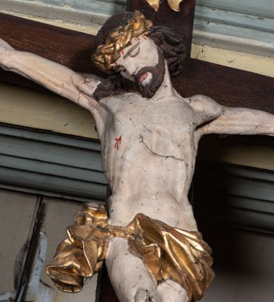 Zdjęcie nr 3: Rzeźbiarska grupa Ukrzyżowania, przedstawiająca przybitego do krzyża Chrystusa podtrzymywanego przez trzy anioły. Figura Chrystusa ścięta z tyłu, przybita do krzyża trzema gwoździami, głowa opadła na prawe ramię, ramiona szeroko rozłożone w delikatnym zwisie. Ciało szczupłe, o wyraźnie podkreślonej anatomii, wydatna klata piersiowa z delikatnie zaznaczoną linią żeber. Twarz o sumarycznie opracowanych rysach, z krótkim, ciemnobrązowym zarostem, dużymi ustami i zamkniętymi oczami. Włosy długie, ciemnobrązowe, falowane, opadające na plecy i prawe ramię grubymi lokami. Na głowie złocona korona cierniowa. Na biodrach krótkie, złocone, przewiązane sznurem perizonium z węzłem i krótkim zwisem na prawym boku. Na czole, dłoniach, stopach i prawym boku delikatne strużki krwi. Kolorystyka ciała naturalistyczna. Krzyż polichromowany na ciemny brąz, nad głową Chrystusa złocony titulus w formie podłużnej banderoli z napisem „IN / RI”. Po prawej stronie, w połowie wysokości dolnej części pionowej belki pełnoplastyczna figura anioła, unoszącego się w powietrzu, z rękami zwróconymi do podtrzymywania krucyfiksu. Głowa w trzech czwartych skierowana w lewo, odchylona do tyłu. Twarz owalna, o wyrazistych rysach, z mocno zaróżowionymi policzkami. Włosy krótkie, kręcone i brązowe. Ubrany jest w złoconą szatę odsłaniającą ramiona, fragment klatki piersiowej i nogi do wysokości kolan. Na plecach ma duże, złocone skrzydła. Belka pozioma flankowana parą przeciwstawnych, unoszących się w powietrzu, pełnoplastycznych putt, podtrzymujących dłońmi krawędzie poziomej belki krzyża. Główki zwrócone frontalnie, twarze o zaokrąglonych, pełnych, wyrazistych rysach i zarumienionych policzkach z dużymi oczami. Włosy krótkie, kręcone i brązowe. Ciała nagie, o zaokrąglonych kształtach, z rozwianymi na plecach, niewielkimi, złoconymi płaszczami i parami małych skrzydełek. Polichromia w odsłoniętych partiach ciała naturalistyczna, szaty i detale złocone.