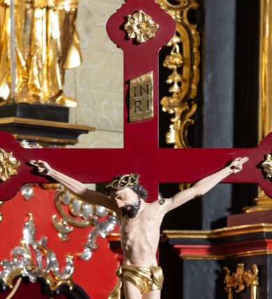Zdjęcie nr 1: Rzeźba pełnoplastyczna, Chrystus przybity do krzyża trzema gwoździami. Figura w delikatnym zwisie, ramiona szeroko rozłożone, kolana delikatnie ugięte, stopy w układzie prawa na lewą. Twarz pociągła, szczupła, o długim nosie, oczy zamknięte, usta szerokie, rozchylone; okolona krótką brodą. Włosy półdługie, ułożone w skręcone pukle opadające na kark i prawe ramię. Na głowie korona cierniowa. Głowa nieproporcjonalnie duża w stosunku do ciała, sylwetka szczupła, z wyraźnie uwypuklonym umięśnieniem i linią żeber klatki piersiowej, z zaznaczonymi śladami męki. Perizonium w formie tkaniny ściśle owijającej biodra, zawiązanej na prawym boku ze zwisem z prawej strony. Nad głową Chrystusa złocony titulus w formie banderoli z napisem: „I(ESUS) N(AZARENUS) / R(EX) I(UDAEROUM)”. Krzyż gładki o trójlistnie zakończonych ramionach, zdobionych rozetkami, malowany na czerwono. Osadzony na podstawie zdobionej dwiema fantazyjnie wyciętymi deseczkami zdobionymi rzeźbionymi, złoconymi gałązkami róż. Podstawa umieszczona na niskim prostokątnym cokoliku na czterech płaskich nóżkach. Polichromia naturalistyczna w odsłoniętych partiach ciała, perizonium i korona złocone.