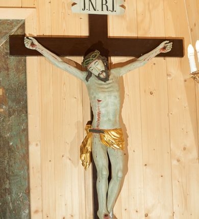 Zdjęcie nr 1: Rzeźba pełna, delikatnie ścięta z tyłu. Figura w typie christo vivo. Ciało w delikatnym zwisie, o szeroko rozłożonych ramionach, delikatnie uniesionych. Nogi lekko ugięte w kolanach, stopy przebite jednym gwoździem, w układzie prawa na lewą. Głowa przechylona na prawe ramię. Twarz pociągła o długim i prostym nosie, oczy wąskie i zmrużone, usta niewielkie; okolona krótką brodą, układającą się w dwa pukle. Włosy długie, silnie skręcone w pasma opadające na kark i prawe ramię. Na głowie zielona korona cierniowa. Ciało plastycznie kształtowane, światłocieniowe, z podkreśloną muskulaturą i ścięgnami, modelowaną w formy guzowate, linią żeber i mostka z zaznaczonymi śladami męki. Perizonium krótkie w postaci tkaniny przewieszonej przez podwójny sznur, odsłaniającej prawe biodro i udo, z kaskadowym zwisem  tkaniny na prawym boku. Na górnej belce krzyża titulus w formie banderoli z napisem „I.N.R.I.”. Polichromia naturalistyczna w odsłoniętych partiach ciała, karnacja bladozielona, perizonium złocone. Krzyż gładki, prosty, malowany na kolor ciemnobrązowy.
