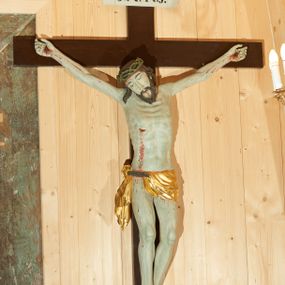 Zdjęcie nr 1: Rzeźba pełna, delikatnie ścięta z tyłu. Figura w typie christo vivo. Ciało w delikatnym zwisie, o szeroko rozłożonych ramionach, delikatnie uniesionych. Nogi lekko ugięte w kolanach, stopy przebite jednym gwoździem, w układzie prawa na lewą. Głowa przechylona na prawe ramię. Twarz pociągła o długim i prostym nosie, oczy wąskie i zmrużone, usta niewielkie; okolona krótką brodą, układającą się w dwa pukle. Włosy długie, silnie skręcone w pasma opadające na kark i prawe ramię. Na głowie zielona korona cierniowa. Ciało plastycznie kształtowane, światłocieniowe, z podkreśloną muskulaturą i ścięgnami, modelowaną w formy guzowate, linią żeber i mostka z zaznaczonymi śladami męki. Perizonium krótkie w postaci tkaniny przewieszonej przez podwójny sznur, odsłaniającej prawe biodro i udo, z kaskadowym zwisem  tkaniny na prawym boku. Na górnej belce krzyża titulus w formie banderoli z napisem „I.N.R.I.”. Polichromia naturalistyczna w odsłoniętych partiach ciała, karnacja bladozielona, perizonium złocone. Krzyż gładki, prosty, malowany na kolor ciemnobrązowy.
