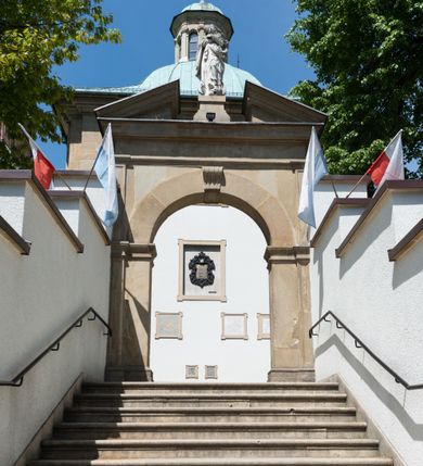 Zdjęcie nr 5: Kościół orientowany, jest budowlą murowaną z cegły i kamienia, na rzucie podłużnym. Korpus jednonawowy i trójprzęsłowy. Stosunkowo szerokie, trójprzęsłowe, zamknięte trójbocznie prezbiterium z przylegającą od północy zakrystią. Na osi kościoła przylega do fasady trójkondygnacyjna wieża z kruchtą w przyziemiu i przyległością od południa, w której mieszczą się prowadzące na wieżę schody.  Od południa i zachodu, na wysokości trzeciego przęsła do korpusu przylega kaplic na rzutach zbliżonych do kwadratów, z czego południową od wschodu i zachodu flankuje para krucht. Nawa, prezbiterium i kaplica północna przekryte są sklepieniami kolebkowymi z lunetami wspartymi na parach lub pojedynczych pilastrach. Kaplica południowa nakryta jest kopułą wspartą na 4 pendentywach. Artykulację ścian na zewnątrz korpusu nawowego i prezbiterium stanowi zespół przypór. Naroża kaplicy południowej ujmują zwielokrotnione pilastry wpierające obiegający bryłę gzyms wieńczący. Naroża kaplicy północnej ujmuje para neogotyckich przypór, natomiast północną ścianę wieńczy neogotycki szczyt schodkowy. 
Do wnętrza prowadzi 6 otworów drzwiowych – 1 w kruchcie w wieży ujęte kamienny, półkoliście zamkniętym portalem schodkowym z dwuskrzydłowymi drzwiami z nadświetlem, 1 zamknięte łukiem półkolistym w przyległości obok wieży, 3 w kruchtach flankujących kaplicę południową – z czego w zachodniej jedno w formie dużych dwuskrzydłowych drzwi z kwaterami bocznymi i nadświetlem, we wschodniej 2 ujęte prostokątnymi, kamiennymi portalami z wąskim nadświetlem i parą uszaków, zwieńczonych parą stykających się wolut z krzyżem na kuli w szczycie oraz 1 prowadzące do zakrystii. Okna korpusu nawowego i prezbiterium w kształcie stojących prostokątów zamkniętych półkoliście, z czego w prezbiterium są smukłe i wysokie. Okna kaplicy północnej zamknięte łukiem koszowym, natomiast kaplicy południowej w postaci prostokątów ujętych kamiennymi obramieniami z uszakami na narożach. 3 z 4 ścian wieży przeprute oknami, po 1 na każdej z kondygnacji, z czego na pierwszej zamknięte półkoliście, na drugiej w kształcie stojącego prostokąta i na trzeciej ostrołukowe. Wieżę wieńczy ceglany szczyt z wydzielonym gzymsami fryzem arkadkowym, wyżej 4 trójkątne szczyty umieszczone na osiach każdej ze ścian ujęte w narożach 4 małymi pinaklami i hełmem pośrodku w formie wysokiego pinakla. Korpus nawowy i prezbiterium przekrywa dach dwuspadowy, kryty blachą, z pinaklową wieżyczką na sygnaturkę na połączeniu tych przestrzeni, kaplicę południową kopuła z latarnią, przylegające do niej kruchty dachy pulpitowe, natomiast kaplica północna ma dach dwuspadowy. Otynkowane ściany kościoła są pomalowane w kolorze białym, miejscami pozostawiony kamienny mur.     
