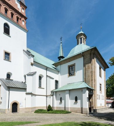 Zdjęcie nr 6: Kościół orientowany, jest budowlą murowaną z cegły i kamienia, na rzucie podłużnym. Korpus jednonawowy i trójprzęsłowy. Stosunkowo szerokie, trójprzęsłowe, zamknięte trójbocznie prezbiterium z przylegającą od północy zakrystią. Na osi kościoła przylega do fasady trójkondygnacyjna wieża z kruchtą w przyziemiu i przyległością od południa, w której mieszczą się prowadzące na wieżę schody.  Od południa i zachodu, na wysokości trzeciego przęsła do korpusu przylega kaplic na rzutach zbliżonych do kwadratów, z czego południową od wschodu i zachodu flankuje para krucht. Nawa, prezbiterium i kaplica północna przekryte są sklepieniami kolebkowymi z lunetami wspartymi na parach lub pojedynczych pilastrach. Kaplica południowa nakryta jest kopułą wspartą na 4 pendentywach. Artykulację ścian na zewnątrz korpusu nawowego i prezbiterium stanowi zespół przypór. Naroża kaplicy południowej ujmują zwielokrotnione pilastry wpierające obiegający bryłę gzyms wieńczący. Naroża kaplicy północnej ujmuje para neogotyckich przypór, natomiast północną ścianę wieńczy neogotycki szczyt schodkowy. 
Do wnętrza prowadzi 6 otworów drzwiowych – 1 w kruchcie w wieży ujęte kamienny, półkoliście zamkniętym portalem schodkowym z dwuskrzydłowymi drzwiami z nadświetlem, 1 zamknięte łukiem półkolistym w przyległości obok wieży, 3 w kruchtach flankujących kaplicę południową – z czego w zachodniej jedno w formie dużych dwuskrzydłowych drzwi z kwaterami bocznymi i nadświetlem, we wschodniej 2 ujęte prostokątnymi, kamiennymi portalami z wąskim nadświetlem i parą uszaków, zwieńczonych parą stykających się wolut z krzyżem na kuli w szczycie oraz 1 prowadzące do zakrystii. Okna korpusu nawowego i prezbiterium w kształcie stojących prostokątów zamkniętych półkoliście, z czego w prezbiterium są smukłe i wysokie. Okna kaplicy północnej zamknięte łukiem koszowym, natomiast kaplicy południowej w postaci prostokątów ujętych kamiennymi obramieniami z uszakami na narożach. 3 z 4 ścian wieży przeprute oknami, po 1 na każdej z kondygnacji, z czego na pierwszej zamknięte półkoliście, na drugiej w kształcie stojącego prostokąta i na trzeciej ostrołukowe. Wieżę wieńczy ceglany szczyt z wydzielonym gzymsami fryzem arkadkowym, wyżej 4 trójkątne szczyty umieszczone na osiach każdej ze ścian ujęte w narożach 4 małymi pinaklami i hełmem pośrodku w formie wysokiego pinakla. Korpus nawowy i prezbiterium przekrywa dach dwuspadowy, kryty blachą, z pinaklową wieżyczką na sygnaturkę na połączeniu tych przestrzeni, kaplicę południową kopuła z latarnią, przylegające do niej kruchty dachy pulpitowe, natomiast kaplica północna ma dach dwuspadowy. Otynkowane ściany kościoła są pomalowane w kolorze białym, miejscami pozostawiony kamienny mur.     
