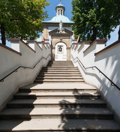 Zdjęcie nr 4: Kościół orientowany, jest budowlą murowaną z cegły i kamienia, na rzucie podłużnym. Korpus jednonawowy i trójprzęsłowy. Stosunkowo szerokie, trójprzęsłowe, zamknięte trójbocznie prezbiterium z przylegającą od północy zakrystią. Na osi kościoła przylega do fasady trójkondygnacyjna wieża z kruchtą w przyziemiu i przyległością od południa, w której mieszczą się prowadzące na wieżę schody.  Od południa i zachodu, na wysokości trzeciego przęsła do korpusu przylega kaplic na rzutach zbliżonych do kwadratów, z czego południową od wschodu i zachodu flankuje para krucht. Nawa, prezbiterium i kaplica północna przekryte są sklepieniami kolebkowymi z lunetami wspartymi na parach lub pojedynczych pilastrach. Kaplica południowa nakryta jest kopułą wspartą na 4 pendentywach. Artykulację ścian na zewnątrz korpusu nawowego i prezbiterium stanowi zespół przypór. Naroża kaplicy południowej ujmują zwielokrotnione pilastry wpierające obiegający bryłę gzyms wieńczący. Naroża kaplicy północnej ujmuje para neogotyckich przypór, natomiast północną ścianę wieńczy neogotycki szczyt schodkowy. 
Do wnętrza prowadzi 6 otworów drzwiowych – 1 w kruchcie w wieży ujęte kamienny, półkoliście zamkniętym portalem schodkowym z dwuskrzydłowymi drzwiami z nadświetlem, 1 zamknięte łukiem półkolistym w przyległości obok wieży, 3 w kruchtach flankujących kaplicę południową – z czego w zachodniej jedno w formie dużych dwuskrzydłowych drzwi z kwaterami bocznymi i nadświetlem, we wschodniej 2 ujęte prostokątnymi, kamiennymi portalami z wąskim nadświetlem i parą uszaków, zwieńczonych parą stykających się wolut z krzyżem na kuli w szczycie oraz 1 prowadzące do zakrystii. Okna korpusu nawowego i prezbiterium w kształcie stojących prostokątów zamkniętych półkoliście, z czego w prezbiterium są smukłe i wysokie. Okna kaplicy północnej zamknięte łukiem koszowym, natomiast kaplicy południowej w postaci prostokątów ujętych kamiennymi obramieniami z uszakami na narożach. 3 z 4 ścian wieży przeprute oknami, po 1 na każdej z kondygnacji, z czego na pierwszej zamknięte półkoliście, na drugiej w kształcie stojącego prostokąta i na trzeciej ostrołukowe. Wieżę wieńczy ceglany szczyt z wydzielonym gzymsami fryzem arkadkowym, wyżej 4 trójkątne szczyty umieszczone na osiach każdej ze ścian ujęte w narożach 4 małymi pinaklami i hełmem pośrodku w formie wysokiego pinakla. Korpus nawowy i prezbiterium przekrywa dach dwuspadowy, kryty blachą, z pinaklową wieżyczką na sygnaturkę na połączeniu tych przestrzeni, kaplicę południową kopuła z latarnią, przylegające do niej kruchty dachy pulpitowe, natomiast kaplica północna ma dach dwuspadowy. Otynkowane ściany kościoła są pomalowane w kolorze białym, miejscami pozostawiony kamienny mur.     
