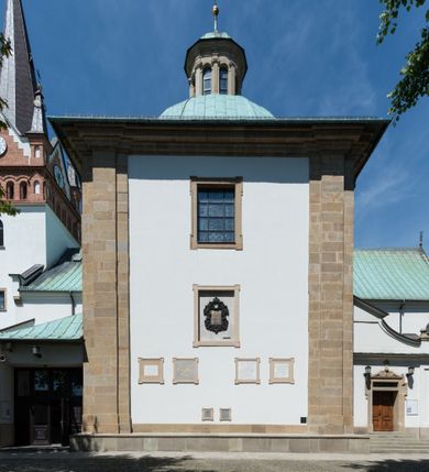 Zdjęcie nr 9: Kościół orientowany, jest budowlą murowaną z cegły i kamienia, na rzucie podłużnym. Korpus jednonawowy i trójprzęsłowy. Stosunkowo szerokie, trójprzęsłowe, zamknięte trójbocznie prezbiterium z przylegającą od północy zakrystią. Na osi kościoła przylega do fasady trójkondygnacyjna wieża z kruchtą w przyziemiu i przyległością od południa, w której mieszczą się prowadzące na wieżę schody.  Od południa i zachodu, na wysokości trzeciego przęsła do korpusu przylega kaplic na rzutach zbliżonych do kwadratów, z czego południową od wschodu i zachodu flankuje para krucht. Nawa, prezbiterium i kaplica północna przekryte są sklepieniami kolebkowymi z lunetami wspartymi na parach lub pojedynczych pilastrach. Kaplica południowa nakryta jest kopułą wspartą na 4 pendentywach. Artykulację ścian na zewnątrz korpusu nawowego i prezbiterium stanowi zespół przypór. Naroża kaplicy południowej ujmują zwielokrotnione pilastry wpierające obiegający bryłę gzyms wieńczący. Naroża kaplicy północnej ujmuje para neogotyckich przypór, natomiast północną ścianę wieńczy neogotycki szczyt schodkowy. 
Do wnętrza prowadzi 6 otworów drzwiowych – 1 w kruchcie w wieży ujęte kamienny, półkoliście zamkniętym portalem schodkowym z dwuskrzydłowymi drzwiami z nadświetlem, 1 zamknięte łukiem półkolistym w przyległości obok wieży, 3 w kruchtach flankujących kaplicę południową – z czego w zachodniej jedno w formie dużych dwuskrzydłowych drzwi z kwaterami bocznymi i nadświetlem, we wschodniej 2 ujęte prostokątnymi, kamiennymi portalami z wąskim nadświetlem i parą uszaków, zwieńczonych parą stykających się wolut z krzyżem na kuli w szczycie oraz 1 prowadzące do zakrystii. Okna korpusu nawowego i prezbiterium w kształcie stojących prostokątów zamkniętych półkoliście, z czego w prezbiterium są smukłe i wysokie. Okna kaplicy północnej zamknięte łukiem koszowym, natomiast kaplicy południowej w postaci prostokątów ujętych kamiennymi obramieniami z uszakami na narożach. 3 z 4 ścian wieży przeprute oknami, po 1 na każdej z kondygnacji, z czego na pierwszej zamknięte półkoliście, na drugiej w kształcie stojącego prostokąta i na trzeciej ostrołukowe. Wieżę wieńczy ceglany szczyt z wydzielonym gzymsami fryzem arkadkowym, wyżej 4 trójkątne szczyty umieszczone na osiach każdej ze ścian ujęte w narożach 4 małymi pinaklami i hełmem pośrodku w formie wysokiego pinakla. Korpus nawowy i prezbiterium przekrywa dach dwuspadowy, kryty blachą, z pinaklową wieżyczką na sygnaturkę na połączeniu tych przestrzeni, kaplicę południową kopuła z latarnią, przylegające do niej kruchty dachy pulpitowe, natomiast kaplica północna ma dach dwuspadowy. Otynkowane ściany kościoła są pomalowane w kolorze białym, miejscami pozostawiony kamienny mur.     
