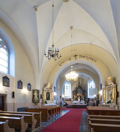 Zdjęcie nr 16: Kościół jest położony w centrum wsi, na południe od głównej drogi, na placu otoczonym płotem na kamiennej podmurówce. Wejście na plac kościelny prowadzi przez główną bramę od zachodu oraz drugą od północnego-wschodu. Kościół jest budowlą założoną na planie krzyża łacińskiego, murowaną, orientowaną z delikatnym odchyleniem ku południowi, jednonawową z transeptem, z prezbiterium zamkniętym trójbocznie, przy którym od północy jest zakrystia z emporą, a od południa kaplica. Wnętrze nawy głównej artykułowe jest dwiema płytkimi wnękami, zakończonymi półkoliście, w których w górnej części umieszczone są trójdzielne okna z maswerkami i witrażami. W prezbiterium i kaplicy sklepienie krzyżowo-żebrowe, w nawie gwieździste bez żeber. Nawę od prezbiterium oraz ramion transeptu oddzielają półkoliście zamknięte arkady. Na ścianie arkady tęczowej napis: „CHWAŁA CI TRÓJCO ŚWIĘTA”. W ramionach transeptu pojedyncze okna prostokątne, zamknięte półkoliście. Kolejne dwa okna, lecz węższe znajdują się w prezbiterium. 

Fasada dwukondygnacyjna z dostawioną wieżą na planie kwadratu. W dolnej kondygnacji fasada artykułowana parą pilastrów w narożach, podtrzymujących profilowany gzyms. Pośrodku główne wejście do kościoła zaakcentowane schodami i monumentalnym portalem z wimpergą i masywnymi kamiennymi węgarami. W górnej kondygnacji fasady uwidoczniona sylwetka wieży z każdej strony flankowana w narożach parą lizen podtrzymujących niepełne belkowanie i trójkątne przyczółki. Od frontu, po bokach wieży spływy wolutowe, w polu półkoliście zamknięte okno, a nad nim okrągłe. Na ścianach bocznych wieży analogiczne oculusy. Na szczycie wieży wysoka iglica. Pozostałe ściany kościoła artykułowe pilastrami, podtrzymującymi niepełne belkowanie zamknięte gzymsem podokapowym. Dach dwuspadowy, nakryty dachówką.

