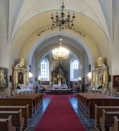 Zdjęcie nr 14: Kościół jest położony w centrum wsi, na południe od głównej drogi, na placu otoczonym płotem na kamiennej podmurówce. Wejście na plac kościelny prowadzi przez główną bramę od zachodu oraz drugą od północnego-wschodu. Kościół jest budowlą założoną na planie krzyża łacińskiego, murowaną, orientowaną z delikatnym odchyleniem ku południowi, jednonawową z transeptem, z prezbiterium zamkniętym trójbocznie, przy którym od północy jest zakrystia z emporą, a od południa kaplica. Wnętrze nawy głównej artykułowe jest dwiema płytkimi wnękami, zakończonymi półkoliście, w których w górnej części umieszczone są trójdzielne okna z maswerkami i witrażami. W prezbiterium i kaplicy sklepienie krzyżowo-żebrowe, w nawie gwieździste bez żeber. Nawę od prezbiterium oraz ramion transeptu oddzielają półkoliście zamknięte arkady. Na ścianie arkady tęczowej napis: „CHWAŁA CI TRÓJCO ŚWIĘTA”. W ramionach transeptu pojedyncze okna prostokątne, zamknięte półkoliście. Kolejne dwa okna, lecz węższe znajdują się w prezbiterium. 

Fasada dwukondygnacyjna z dostawioną wieżą na planie kwadratu. W dolnej kondygnacji fasada artykułowana parą pilastrów w narożach, podtrzymujących profilowany gzyms. Pośrodku główne wejście do kościoła zaakcentowane schodami i monumentalnym portalem z wimpergą i masywnymi kamiennymi węgarami. W górnej kondygnacji fasady uwidoczniona sylwetka wieży z każdej strony flankowana w narożach parą lizen podtrzymujących niepełne belkowanie i trójkątne przyczółki. Od frontu, po bokach wieży spływy wolutowe, w polu półkoliście zamknięte okno, a nad nim okrągłe. Na ścianach bocznych wieży analogiczne oculusy. Na szczycie wieży wysoka iglica. Pozostałe ściany kościoła artykułowe pilastrami, podtrzymującymi niepełne belkowanie zamknięte gzymsem podokapowym. Dach dwuspadowy, nakryty dachówką.

