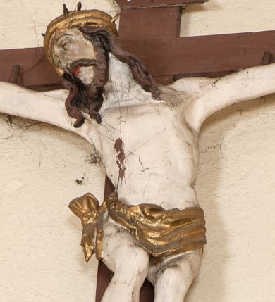 Zdjęcie nr 1: Rzeźba pełna. Figura Chrystusa ukazanego w niewielkim zwisie, o szeroko rozłożonych, niemal prostych ramionach, głowa uniesiona i przechylona na prawy bok, nogi ugięte w kolanach, stopy przebite jednym gwoździem w układzie prawa na lewą. Twarz pociągła, okolona zarostem, z długim i wąskim nosem, z częściowo otwartymi oczami. Włosy długie opadające na ramiona w postaci silnie skręconych pukli. Ciało o niewłaściwych proporcjach, umownie zarysowanej klatce piersiowej. Na głowie korona cierniowa w formie kołpaka. Perizonium z pasa tkaniny, krótkie, obficie i płasko drapowane, zawieszone na sznurze, z końcem przewiązanym na prawym boku. Polichromia karnacji widocznej w odsłoniętych partiach ciała naturalistyczna, korona i perizonium złocone. Na zakończeniu górnej belki krzyża titulus z napisem „INRI”, białym na brązowym tle. Krzyż prosty malowany na kolor brązowy. 