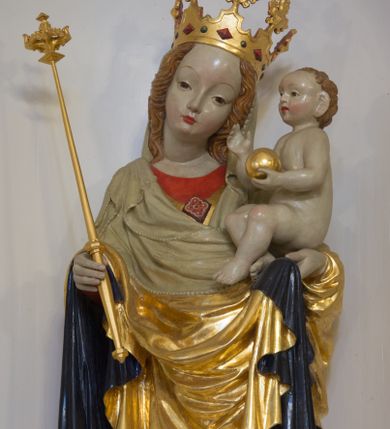 Zdjęcie nr 1: Rzeźba pełnoplastyczna, wolnostojąca, ukazująca Matkę Boską z Dzieciątkiem. Maria ustawiona frontalnie, w kontrapoście o sylwetce wygiętej na kształt litery „S”, z głową przechyloną na lewo. Na lewym ręku trzyma Dzieciątko i podtrzymuje dłonią płaszcz, w prawej dzierży berło. Twarz okrągła z wysokim czołem, drobnymi ustami i małymi oczami, o delikatnie zarysowanych łukach brwiowych. Włosy jasne, ułożone w skręcone pukle spływające na plecy. Madonna ubrana jest w czerwoną suknię, złoty płaszcz z ciemnogranatową podszewką spięty na piersi broszą w kształcie rombu oraz czarne buty. Na głowie ma założony beżowy welon spływający na ramiona i piersi oraz złotą koronę w typie otwartym, kameryzowaną. Draperia szat miękka o wyraźnie zaznaczonych, licznych fałdach misowych i kaskadowych. Dzieciątko ukazane w pozycji siedzącej, zwrócone w prawo, z nóżkami skrzyżowanymi, założonymi lewa na prawą. W lewej dłoni trzyma jabłko królewskie, a prawą podnosi do gestu błogosławieństwa. Twarz o dziecięcych rysach, z wysokim i wypukłym czołem, z delikatnie otwartymi ustami, okolona krótkimi i kędzierzawymi włosami. Polichromia w odsłoniętych partiach ciała o delikatnych odcieniach szarości, płaszcz i atrybuty złocone. 