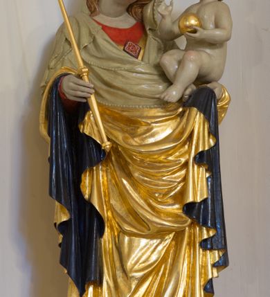Zdjęcie nr 2: Rzeźba pełnoplastyczna, wolnostojąca, ukazująca Matkę Boską z Dzieciątkiem. Maria ustawiona frontalnie, w kontrapoście o sylwetce wygiętej na kształt litery „S”, z głową przechyloną na lewo. Na lewym ręku trzyma Dzieciątko i podtrzymuje dłonią płaszcz, w prawej dzierży berło. Twarz okrągła z wysokim czołem, drobnymi ustami i małymi oczami, o delikatnie zarysowanych łukach brwiowych. Włosy jasne, ułożone w skręcone pukle spływające na plecy. Madonna ubrana jest w czerwoną suknię, złoty płaszcz z ciemnogranatową podszewką spięty na piersi broszą w kształcie rombu oraz czarne buty. Na głowie ma założony beżowy welon spływający na ramiona i piersi oraz złotą koronę w typie otwartym, kameryzowaną. Draperia szat miękka o wyraźnie zaznaczonych, licznych fałdach misowych i kaskadowych. Dzieciątko ukazane w pozycji siedzącej, zwrócone w prawo, z nóżkami skrzyżowanymi, założonymi lewa na prawą. W lewej dłoni trzyma jabłko królewskie, a prawą podnosi do gestu błogosławieństwa. Twarz o dziecięcych rysach, z wysokim i wypukłym czołem, z delikatnie otwartymi ustami, okolona krótkimi i kędzierzawymi włosami. Polichromia w odsłoniętych partiach ciała o delikatnych odcieniach szarości, płaszcz i atrybuty złocone. 