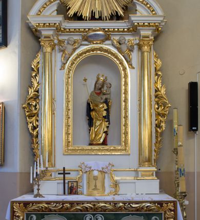 Zdjęcie nr 9: Rzeźba pełnoplastyczna, wolnostojąca, ukazująca Matkę Boską z Dzieciątkiem. Maria ustawiona frontalnie, w kontrapoście o sylwetce wygiętej na kształt litery „S”, z głową przechyloną na lewo. Na lewym ręku trzyma Dzieciątko i podtrzymuje dłonią płaszcz, w prawej dzierży berło. Twarz okrągła z wysokim czołem, drobnymi ustami i małymi oczami, o delikatnie zarysowanych łukach brwiowych. Włosy jasne, ułożone w skręcone pukle spływające na plecy. Madonna ubrana jest w czerwoną suknię, złoty płaszcz z ciemnogranatową podszewką spięty na piersi broszą w kształcie rombu oraz czarne buty. Na głowie ma założony beżowy welon spływający na ramiona i piersi oraz złotą koronę w typie otwartym, kameryzowaną. Draperia szat miękka o wyraźnie zaznaczonych, licznych fałdach misowych i kaskadowych. Dzieciątko ukazane w pozycji siedzącej, zwrócone w prawo, z nóżkami skrzyżowanymi, założonymi lewa na prawą. W lewej dłoni trzyma jabłko królewskie, a prawą podnosi do gestu błogosławieństwa. Twarz o dziecięcych rysach, z wysokim i wypukłym czołem, z delikatnie otwartymi ustami, okolona krótkimi i kędzierzawymi włosami. Polichromia w odsłoniętych partiach ciała o delikatnych odcieniach szarości, płaszcz i atrybuty złocone. 