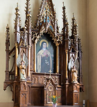 Zdjęcie nr 1: Ołtarz drewniany, prostopadłościenny, na dwustopniowej podstawie; mensa drewniana. Antepedium ujęte w narożach parą kolumienek, pole podzielone na trzy płyciny z maswerkami: zewnętrzne prostokątne, środkowa kwadratowa z monogramem Marii. Tabernakulum w kształcie drewnianej prostopadłościennej skrzynki, w formie kapliczki. Flankowane od frontu dwiema lizenami zakończonymi sterczynami z żabkami; nad polem środkowym zamknięte wimpergą. Drzwiczki w formie stojącego prostokąta zamkniętego łukiem ostrym, dekorowane w polu dwiema blendami i rozetą. Nastawa ołtarzowa architektoniczna, jednoosiowa, jednokondygnacyjna ze zwieńczeniem, z wysoką predellą. Kondygnacja ujęta dwiema parami kolumienek na wysokich, wąskich cokołach, dźwigających podwójne sterczyny, pomiędzy nimi cokoły na figury aniołów, zwieńczone baldachimami utworzonymi z konstrukcji dwóch łuków ostrych i sterczyny. W polu środkowym nisza w formie stojącego prostokąta zamkniętego łukiem ostrym, ujęta kolumienkami o skręconych trzonach (w dolnej części trzony gładkie), z obrazem św. Teresy z Lisieux; w przyłuczach dekorowana maswerkiem; zamknięta baldachimem utworzonym naprzemiennie z łuków ostrych i sterczyn. Kondygnacja ujęta po zewnętrznej stronie przyporami podtrzymywanymi przez wsporniki. Zwieńczenie w formie wimpergi dekorowanej żabkami, zakończonej krzyżem; dekorowanej maswerkiem w polu. Struktura w kolorze naturalnego drewna; detale w kolorze niebieskim, kolumny, dekoracja i profile złocone.
