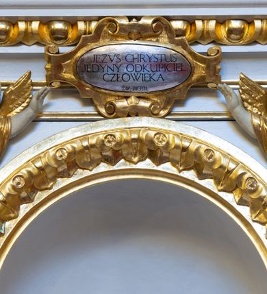 Zdjęcie nr 10: Ołtarz drewniany, prostopadłościenny, na jednostopniowej podstawie. Mensa drewniana z portatylem z marmuru dębnickiego z kwadratowym reservaculum na relikwie św. Konstancji i św. Teodora. Antepedium w formie leżącego  prostokąta ujętego grubą ramą, dekorowaną płaskorzeźbionym akantem. W polu, w centrum malowany na desce rollwerk z monogramem Marii w polu, ujęty wolutami utworzonymi z ornamentu roślinnego o wici zakończonej kampanulą. Tabernakulum w formie prostopadłościennej skrzynki o delikatnie wypukłej ściance frontowej. Całość zamknięta opasującym je z trzech stron gzymsem przyjmującym nad polem środkowym kształt łuku segmentowego podtrzymywanego przez dwie konsole utworzone z akantu. Drzwiczki w kształcie stojącego prostokąta zamkniętego łukiem półkolistym nadwieszonym, ujęte ramą. W polu ryte przedstawienie kielicha z hostią. Tabernakulum flankowane spływami wolutowymi utworzonymi z akantu. Nastawa ołtarzowa architektoniczna, jednoosiowa, jednokondygnacyjna ze zwieńczeniem, na niewielkim cokole. Kondygnacja ujęta parą kolumn o kapitelach kompozytowych dźwigających pełne belkowanie wyłamane na ich osi oraz przerwany półkolisty przyczółek. W polu środkowym nisza w formie stojącego prostokąta zamkniętego łukiem półkolistym, ujęta ramą z figurą Matki Boskiej z Dzieciątkiem. Zwieńczenie w formie medalionu z płaskorzeźbionym monogramem Marii w glorii promienistej. W przyłuczach niszy w polu środkowym para puttów podtrzymujących owalny kartusz z napisem w polu „WIELBIJ / DUSZO MOJA PANA / ŚW(IĘTY) ŁUKASZ I R(OZDZIAŁ)”. Kondygnacja flankowana uszami utworzonymi z suchego akantu. Gzyms oraz przerwany przyczółek dekorowane kimationem. Struktura polichromowana w kolorze białym, ornamentyka, profile i kolumny złocone.
