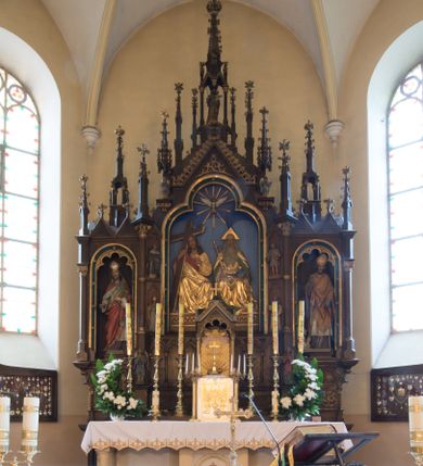 Zdjęcie nr 1: Nastawa ołtarzowa architektoniczna, jednokondygnacyjna, trójosiowa. Ołtarz kamienny, prostopadłościenny, ustawiony na dwustopniowej podstawie. Mensa kamienna, silnie wysunięta, podtrzymywana przez dwie kamienne kolumienki. Antepedium podzielone na trzy kwadratowe pola z płycinami, w środkowej wysuniętej do przodu medalion z monogramem IHS w polu. Tabernakulum w formie prostopadłościennej skrzynki, obitej fakturowaną blachą, od frontu ujęte parą kolumienek. Drzwiczki w kształcie stojącego prostokąta, dwuskrzydłowe z trybowanym przedstawieniem kielicha z unoszącą się Hostią w glorii, adorowaną przez dwa klęczące anioły. Tron do wystawiania monstrancji w formie baldachimu podtrzymywanego przez cztery kolumienki oraz z zabudowaną ścianką tylną dekorowaną rytym wzorem z czteroliści i medalionu z monogramem IHS w glorii podtrzymywanym przez parę unoszących się aniołów. Baldachim w formie dwuspadowego daszku zwieńczonego pseudowieżyczką z krzyżem; od frontu zamknięty wimpergą. Nastawa ołtarzowa ustawiona na dwustrefowym cokole z predellą dekorowaną blendami. Osie wydzielone czterema kolumnami dźwigającymi pinakle. Oś środkowa, najszersza ujęta dwiema dwustrefowymi płytkimi niszami zakończonymi trójlistnie z półpełnymi figurkami świętych: Dominika i Izydora po prawej oraz Ambrożego i Jacka po lewej; zwieńczona wimpergą, flankowaną przez dwie figurki aniołków nakryte baldachimami w formie sterczyn. W polu nisza w formie stojącego prostokąta zamkniętego łukiem trójlistnym z płaskorzeźbionym przedstawieniem Trójcy Świętej. Osie boczne z niszami w formie stojącego prostokąta zamkniętego łukiem półkolistym z figurami św. Wojciecha (po lewej) i św. Stanisława (po prawej), zamknięte dwiema wimpergami ze sterczynami oraz jednospadowym daszkiem z ażurową wieżyczką. Zwieńczenie w formie ażurowej wieżyczki z figurką św. Józefa z Dzieciątkiem, zakończonej dużą sterczyną z krzyżem dekorowaną żabkami. Struktura w kolorze naturalnego drewna, profile i ornamentyka złocone, tło pola środkowego w kolorze błękitnym, detale zielone.
