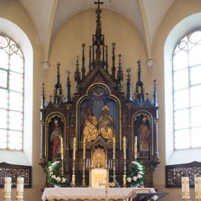 Zdjęcie nr 1: Nastawa ołtarzowa architektoniczna, jednokondygnacyjna, trójosiowa. Ołtarz kamienny, prostopadłościenny, ustawiony na dwustopniowej podstawie. Mensa kamienna, silnie wysunięta, podtrzymywana przez dwie kamienne kolumienki. Antepedium podzielone na trzy kwadratowe pola z płycinami, w środkowej wysuniętej do przodu medalion z monogramem IHS w polu. Tabernakulum w formie prostopadłościennej skrzynki, obitej fakturowaną blachą, od frontu ujęte parą kolumienek. Drzwiczki w kształcie stojącego prostokąta, dwuskrzydłowe z trybowanym przedstawieniem kielicha z unoszącą się Hostią w glorii, adorowaną przez dwa klęczące anioły. Tron do wystawiania monstrancji w formie baldachimu podtrzymywanego przez cztery kolumienki oraz z zabudowaną ścianką tylną dekorowaną rytym wzorem z czteroliści i medalionu z monogramem IHS w glorii podtrzymywanym przez parę unoszących się aniołów. Baldachim w formie dwuspadowego daszku zwieńczonego pseudowieżyczką z krzyżem; od frontu zamknięty wimpergą. Nastawa ołtarzowa ustawiona na dwustrefowym cokole z predellą dekorowaną blendami. Osie wydzielone czterema kolumnami dźwigającymi pinakle. Oś środkowa, najszersza ujęta dwiema dwustrefowymi płytkimi niszami zakończonymi trójlistnie z półpełnymi figurkami świętych: Dominika i Izydora po prawej oraz Ambrożego i Jacka po lewej; zwieńczona wimpergą, flankowaną przez dwie figurki aniołków nakryte baldachimami w formie sterczyn. W polu nisza w formie stojącego prostokąta zamkniętego łukiem trójlistnym z płaskorzeźbionym przedstawieniem Trójcy Świętej. Osie boczne z niszami w formie stojącego prostokąta zamkniętego łukiem półkolistym z figurami św. Wojciecha (po lewej) i św. Stanisława (po prawej), zamknięte dwiema wimpergami ze sterczynami oraz jednospadowym daszkiem z ażurową wieżyczką. Zwieńczenie w formie ażurowej wieżyczki z figurką św. Józefa z Dzieciątkiem, zakończonej dużą sterczyną z krzyżem dekorowaną żabkami. Struktura w kolorze naturalnego drewna, profile i ornamentyka złocone, tło pola środkowego w kolorze błękitnym, detale zielone.
