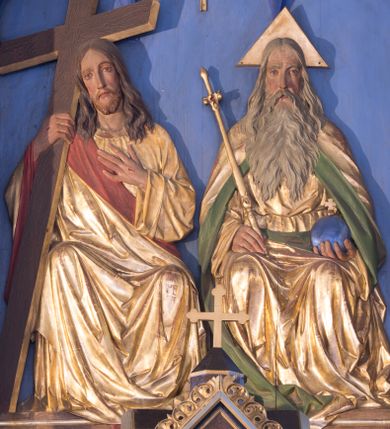 Zdjęcie nr 1: Płaskorzeźbiona grupa przedstawiająca Boga Ojca, Jezusa Chrystusa i Ducha Świętego ustawiona w trójkonchowej niszy. Po prawej stronie Bóg Ojciec ukazany frontalnie, w pozycji siedzącej. W prawej ręce trzyma złote berło, a w lewej niebieskie jabłko zwieńczone  złotym, równoramiennym krzyżykiem. Twarz podłużna z długim i wąskim nosem, małymi ustami, dużymi, błękitnymi oczami, okolona długą, siwą i bujną brodą. Włosy siwe, długie, z przedziałkiem na środku głowy, opadające na ramiona i plecy.  Bóg Ojciec ubrany jest w długą, złotą szatę z długimi rękawami, przepasaną w  pasie, silnie drapowaną oraz złoty płaszcz z zieloną podszewką odwiniętą po bokach, opadający na plecy i dołem z prawej strony. Wokół głowy ma złoty, trójkątny nimb. Po lewej stronie Chrystus ukazany frontalnie, w pozycji siedzącej, w prawej ręce trzyma prosty, brązowy krzyż o złoconych krawędziach; lewą rękę ma złożoną na piersi; głowa delikatnie zwrócona w prawo. Twarz pociągła z długim i wąskim nosem oraz małymi ustami, okolona krótką i brązową brodą. Włosy długie, brązowe, z przedziałkiem na środku głowy, opadające na ramiona i plecy. Chrystus ubrany jest w złotą suknię z długimi rękawami oraz złoty płaszcz z czerwoną podszewką, przerzucony przez prawe ramię i spływający na lewy bok; tkanina silnie drapowana. Na dłoniach ma widoczne ślady po gwoździach. U góry grupy znajduje się gołębica Ducha Świętego  z rozpostartymi szeroko skrzydłami umieszczona w otoku złotych promieni, równolegle ułożonych. Tło jednolite, niebieskie, u dołu podłużna płycina zdobiona gęsto ułożoną, złoconą linią na czerwonym tle. Polichromia w odsłoniętych partiach ciała naturalistyczna.
