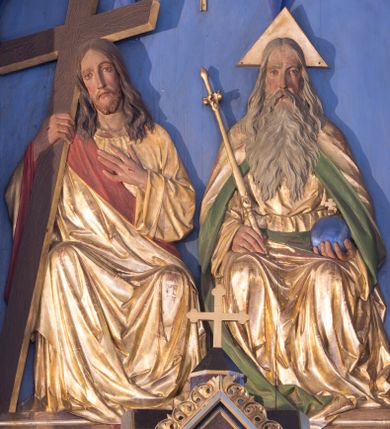 Zdjęcie nr 1: Płaskorzeźbiona grupa przedstawiająca Boga Ojca, Jezusa Chrystusa i Ducha Świętego ustawiona w trójkonchowej niszy. Po prawej stronie Bóg Ojciec ukazany frontalnie, w pozycji siedzącej. W prawej ręce trzyma złote berło, a w lewej niebieskie jabłko zwieńczone  złotym, równoramiennym krzyżykiem. Twarz podłużna z długim i wąskim nosem, małymi ustami, dużymi, błękitnymi oczami, okolona długą, siwą i bujną brodą. Włosy siwe, długie, z przedziałkiem na środku głowy, opadające na ramiona i plecy.  Bóg Ojciec ubrany jest w długą, złotą szatę z długimi rękawami, przepasaną w  pasie, silnie drapowaną oraz złoty płaszcz z zieloną podszewką odwiniętą po bokach, opadający na plecy i dołem z prawej strony. Wokół głowy ma złoty, trójkątny nimb. Po lewej stronie Chrystus ukazany frontalnie, w pozycji siedzącej, w prawej ręce trzyma prosty, brązowy krzyż o złoconych krawędziach; lewą rękę ma złożoną na piersi; głowa delikatnie zwrócona w prawo. Twarz pociągła z długim i wąskim nosem oraz małymi ustami, okolona krótką i brązową brodą. Włosy długie, brązowe, z przedziałkiem na środku głowy, opadające na ramiona i plecy. Chrystus ubrany jest w złotą suknię z długimi rękawami oraz złoty płaszcz z czerwoną podszewką, przerzucony przez prawe ramię i spływający na lewy bok; tkanina silnie drapowana. Na dłoniach ma widoczne ślady po gwoździach. U góry grupy znajduje się gołębica Ducha Świętego  z rozpostartymi szeroko skrzydłami umieszczona w otoku złotych promieni, równolegle ułożonych. Tło jednolite, niebieskie, u dołu podłużna płycina zdobiona gęsto ułożoną, złoconą linią na czerwonym tle. Polichromia w odsłoniętych partiach ciała naturalistyczna.
