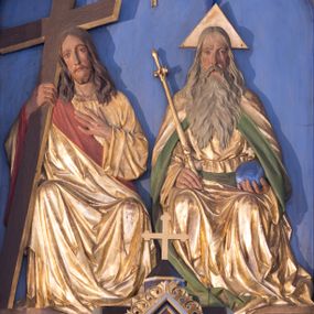 Zdjęcie nr 1: Płaskorzeźbiona grupa przedstawiająca Boga Ojca, Jezusa Chrystusa i Ducha Świętego ustawiona w trójkonchowej niszy. Po prawej stronie Bóg Ojciec ukazany frontalnie, w pozycji siedzącej. W prawej ręce trzyma złote berło, a w lewej niebieskie jabłko zwieńczone  złotym, równoramiennym krzyżykiem. Twarz podłużna z długim i wąskim nosem, małymi ustami, dużymi, błękitnymi oczami, okolona długą, siwą i bujną brodą. Włosy siwe, długie, z przedziałkiem na środku głowy, opadające na ramiona i plecy.  Bóg Ojciec ubrany jest w długą, złotą szatę z długimi rękawami, przepasaną w  pasie, silnie drapowaną oraz złoty płaszcz z zieloną podszewką odwiniętą po bokach, opadający na plecy i dołem z prawej strony. Wokół głowy ma złoty, trójkątny nimb. Po lewej stronie Chrystus ukazany frontalnie, w pozycji siedzącej, w prawej ręce trzyma prosty, brązowy krzyż o złoconych krawędziach; lewą rękę ma złożoną na piersi; głowa delikatnie zwrócona w prawo. Twarz pociągła z długim i wąskim nosem oraz małymi ustami, okolona krótką i brązową brodą. Włosy długie, brązowe, z przedziałkiem na środku głowy, opadające na ramiona i plecy. Chrystus ubrany jest w złotą suknię z długimi rękawami oraz złoty płaszcz z czerwoną podszewką, przerzucony przez prawe ramię i spływający na lewy bok; tkanina silnie drapowana. Na dłoniach ma widoczne ślady po gwoździach. U góry grupy znajduje się gołębica Ducha Świętego  z rozpostartymi szeroko skrzydłami umieszczona w otoku złotych promieni, równolegle ułożonych. Tło jednolite, niebieskie, u dołu podłużna płycina zdobiona gęsto ułożoną, złoconą linią na czerwonym tle. Polichromia w odsłoniętych partiach ciała naturalistyczna.
